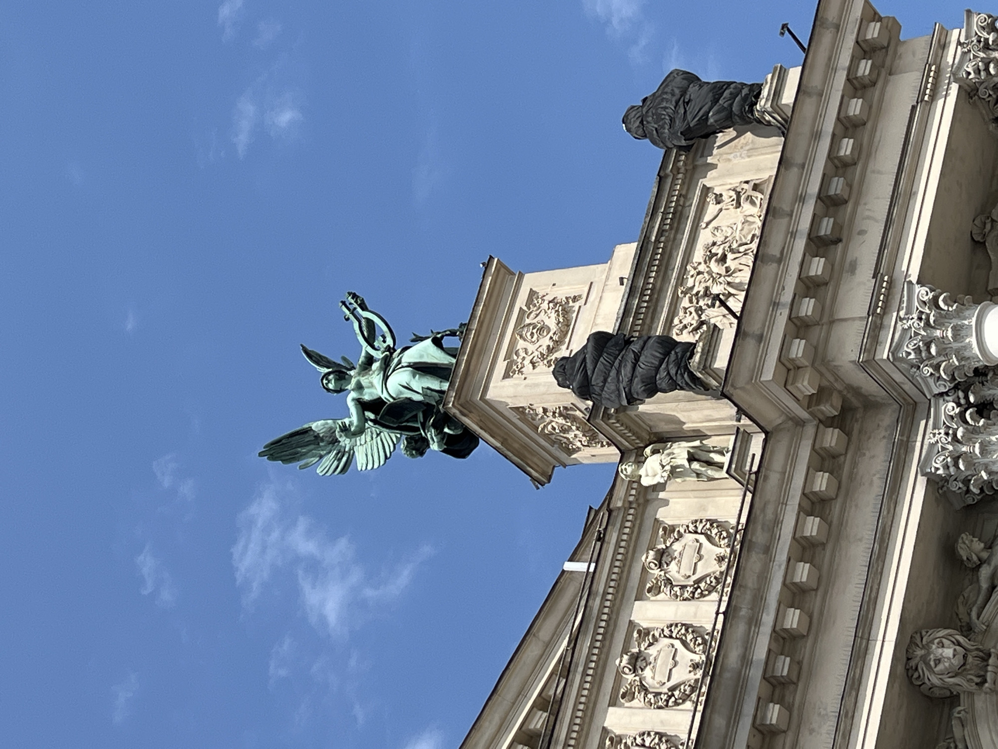 This is a low-angle, outdoor, daytime photograph of the Solomiya Krushelnytska Lviv State Academic Theatre of Opera and Ballet in Lviv, Ukraine. The image is dominated by a bright blue sky with scattered white clouds. The top of the opera house is visible, featuring intricate architectural details. At the apex of the structure is a large, bronze statue of an angel with outstretched wings, holding a lyre and a wreath. Below the angel, on the roofline of the opera house, are several sculptures. On the right and left sides of the roof, there are two dark, somewhat abstract-looking statues. In the center, between these dark statues and closer to the angel sculpture, are two ornate relief panels. Below these, on a lower level of the building, there are further relief sculptures and decorative elements, including what appears to be a lion's head carving. A small, pale statue of a human figure stands on the ledge between the dark statues and the relief panels. The building's facade is made of light-colored stone, and the overall impression is one of grandeur and classical design.