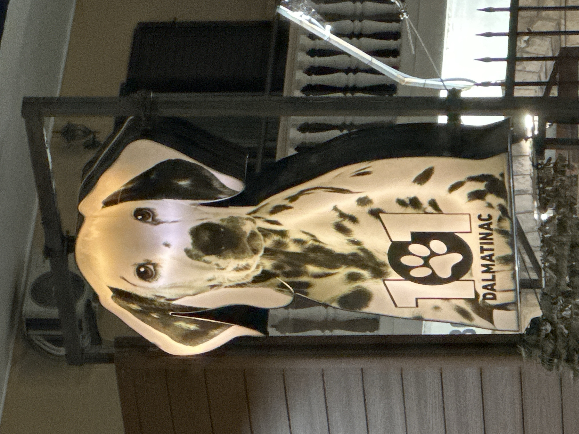 The image shows a sign for "Dalmatinac," a business located in Donji Pobori, Montenegro. The sign features the head of a Dalmatian dog, alongside a paw print within the number 101. The sign is mounted on a metal frame. The backdrop includes a view of a building with a balcony and fence.