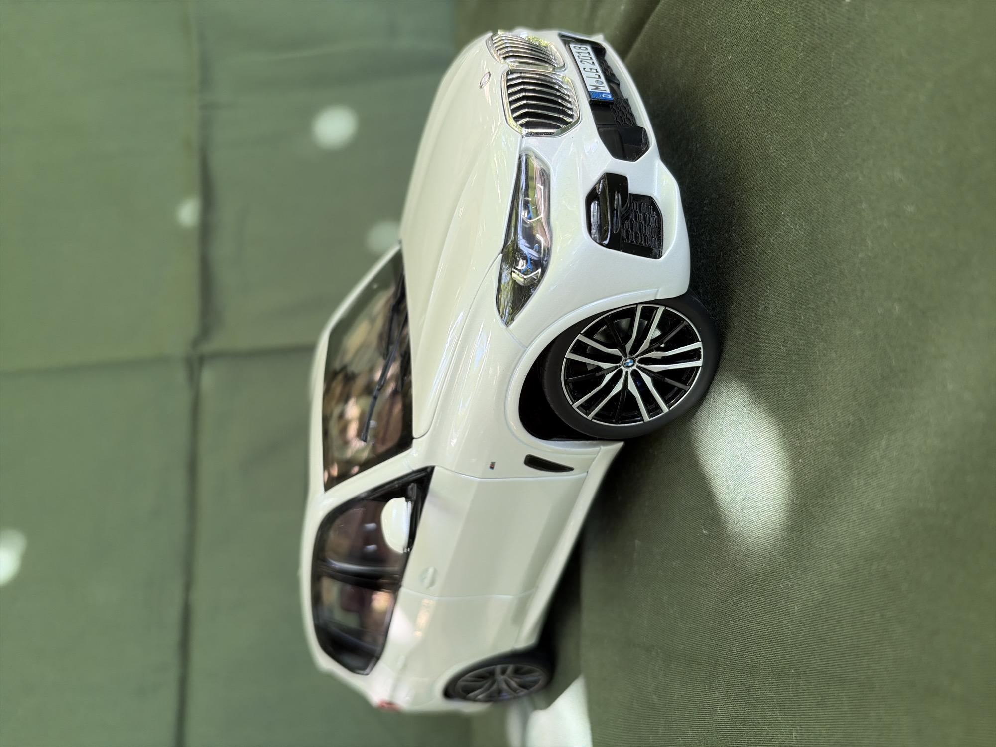 A white BMW X5 model car is parked on a textured green surface. The car is viewed from a low angle, with the front right of the vehicle closest to the viewer. The car's headlights are illuminated. A license plate on the front bumper reads "M LG 2018". The background is a blurred green surface with a series of horizontal lines, suggesting a quilted or upholstered material. There are a few soft, white highlights on the background, possibly from light sources. The scene is static and takes place indoors or in a sheltered outdoor area. The location is not specified as Oxentea, Moldova, as this information is not present in the image.