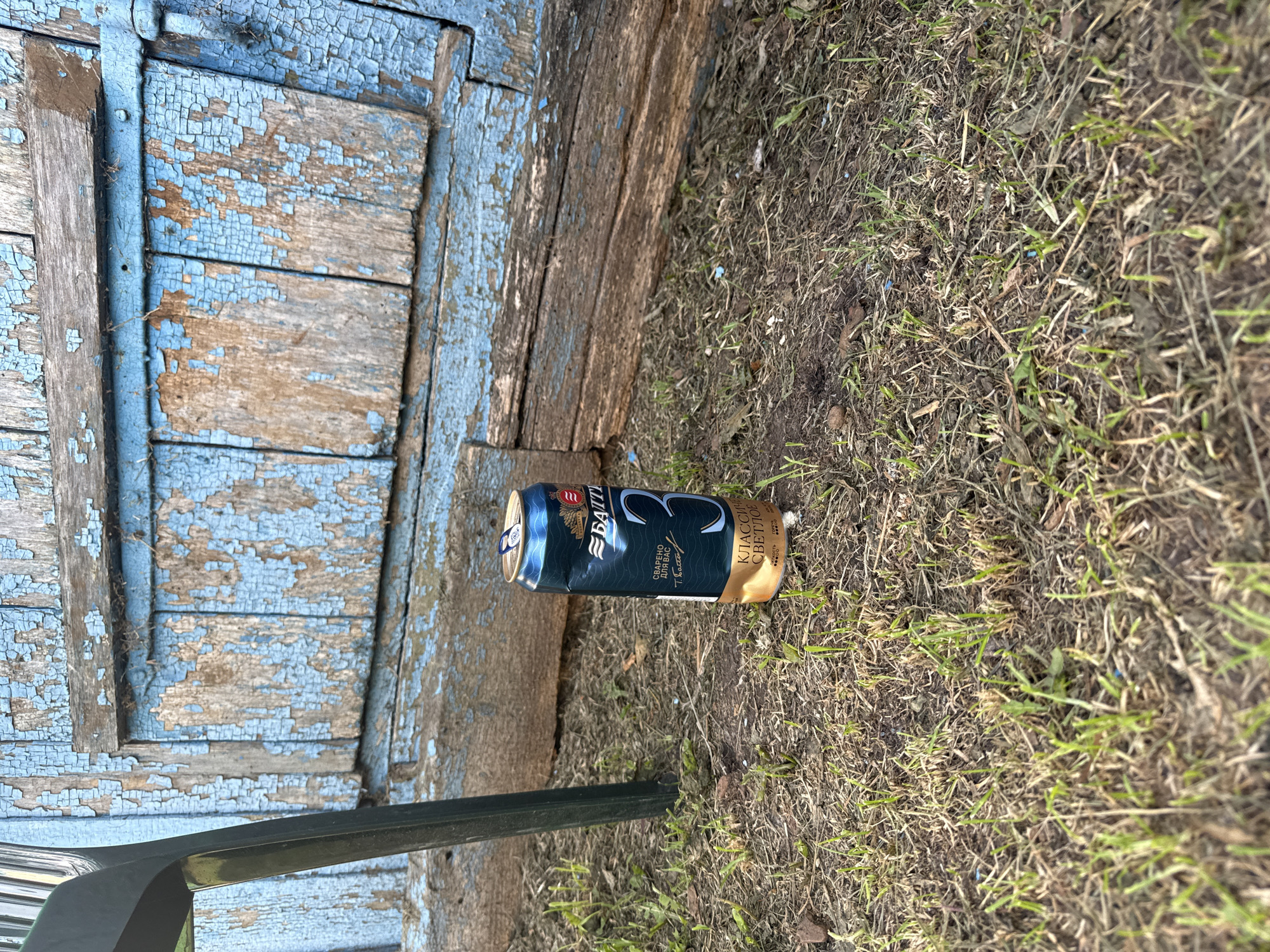 A single, unopened can of beer, specifically "Battle #3" brand, sits upright on dry grass and dirt. The can is primarily dark green with gold and white lettering, featuring a large "3" on its side. Behind the can, a weathered, light blue wooden structure with peeling paint and visible grain dominates the background. A dark green chair leg is visible on the left side of the frame. The overall environment appears to be outdoors, possibly in a rural or neglected urban setting. The location is identified as Chuya-Ataevo, Russia.
