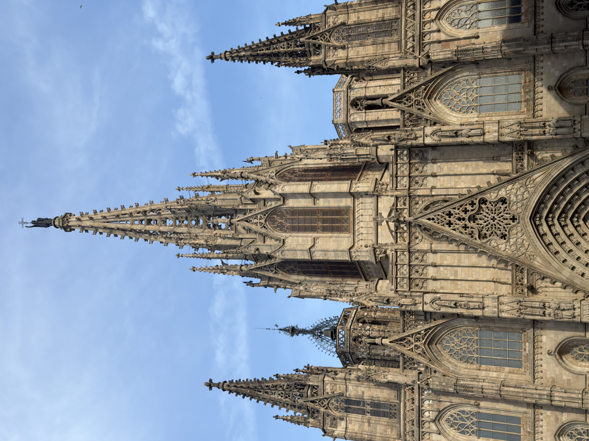 The image shows a detailed view of the Barcelona Cathedral's facade, a Gothic cathedral located in the Barri Gòtic (Gothic Quarter) of the city of Barcelona, Spain. The cathedral features intricate stone carvings, pointed arches, and multiple spires reaching towards a blue sky with scattered clouds. The central spire is topped with a statue and a cross. Other architectural elements visible include large stained-glass windows and decorative stonework, characteristic of Gothic architecture. The lighting suggests it is daytime, likely morning or afternoon. There are no people or discernible actions in the image, focusing solely on the architectural grandeur of the cathedral.