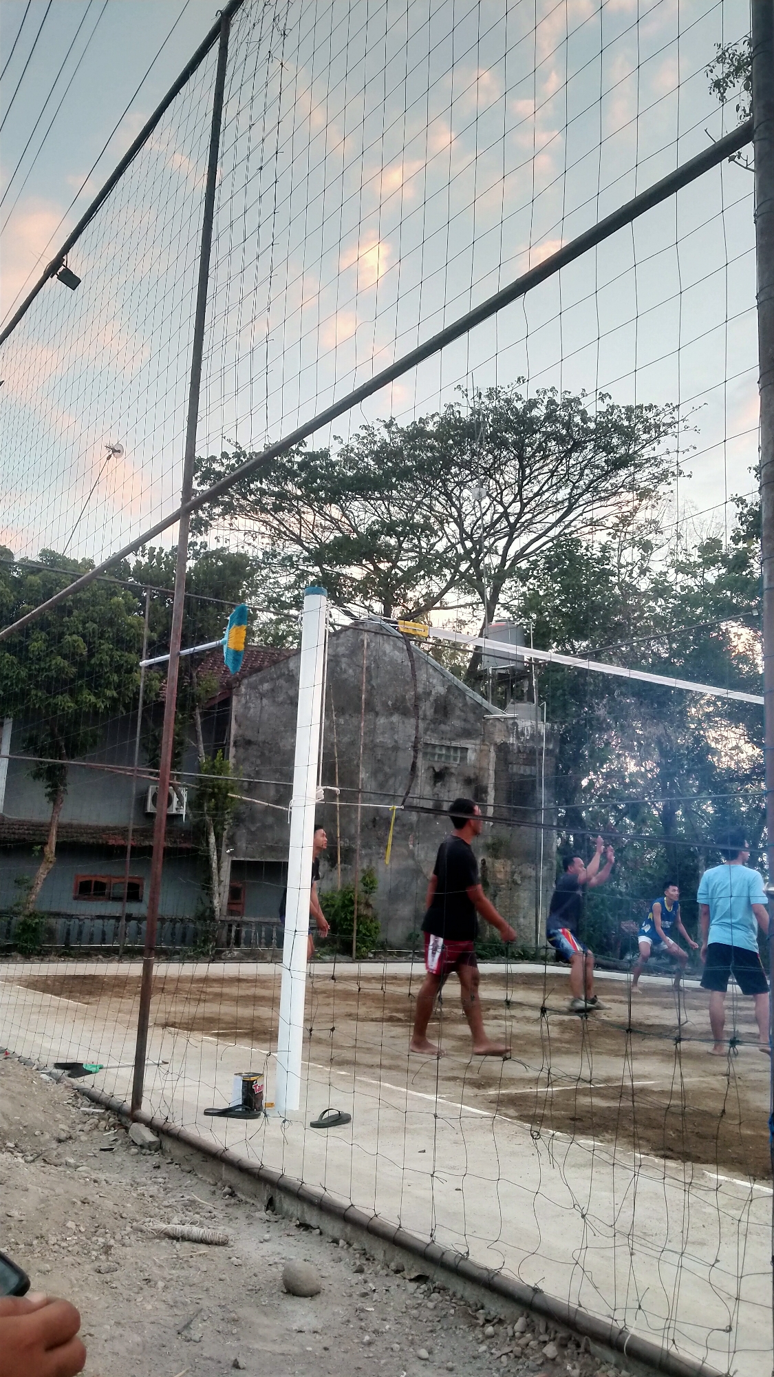 This is a vertical, eye-level, full-shot photograph of an outdoor volleyball game at dusk in Ketoyan, Indonesia. The image is taken from behind a volleyball net, looking towards a volleyball court. The court itself is made of a light-colored, packed dirt surface with visible lines demarcating the playing area. A white volleyball net is strung across the middle of the court, supported by white poles.
There are at least five individuals on the court. On the left side of the net, a person in a black shirt and red and white shorts is in motion, facing away from the camera, appearing to be running or walking. To their right, further back, a person in a dark blue shirt and patterned shorts is captured mid-air, in the act of hitting a volleyball. In the background, another person in a light blue shirt and dark shorts stands facing towards the net. A fourth person in a yellow and blue jersey is visible further back on the court. The foreground shows a rough, dirt and gravel ground with a few scattered stones and a pair of black flip-flops.
Behind the court, a two-story concrete building with visible windows and an air conditioning unit on the side can be seen. Several trees with lush green foliage surround the building, and larger, more mature trees with bare branches are visible in the background against a soft, clouded sky with hints of orange and pink from the setting sun. The overall impression is a casual game of volleyball in a semi-urban or rural setting as daylight fades.