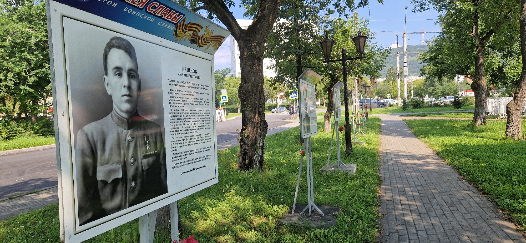 The image shows a memorial display in Miass, Russia, likely dedicated to local heroes. In the foreground, a large portrait of a man in military uniform is mounted on a white board, accompanied by text providing biographical information. The portrait is of Mikhail Petrovich Kushnov, who was born in 1923 in Novotroitsk, Chelyabinsk Oblast, and died in combat in 1944. The text details his military service, including his participation in the Battle of Stalingrad and his actions on the Dnieper River, for which he was posthumously awarded the Hero of the Soviet Union.
The display is part of a larger outdoor exhibition, with several similar boards visible further down a paved walkway. Red artificial poppies are placed at the base of some of these boards, suggesting a commemoration of fallen soldiers, possibly for Victory Day. The exhibition is situated on a grassy area alongside a road and sidewalk, with trees providing shade. In the background, buildings and a distant industrial structure with a red and white tower are visible, indicating an urban or suburban setting within the city of Miass. The weather appears clear and sunny, with shadows cast by the trees and the memorial boards.