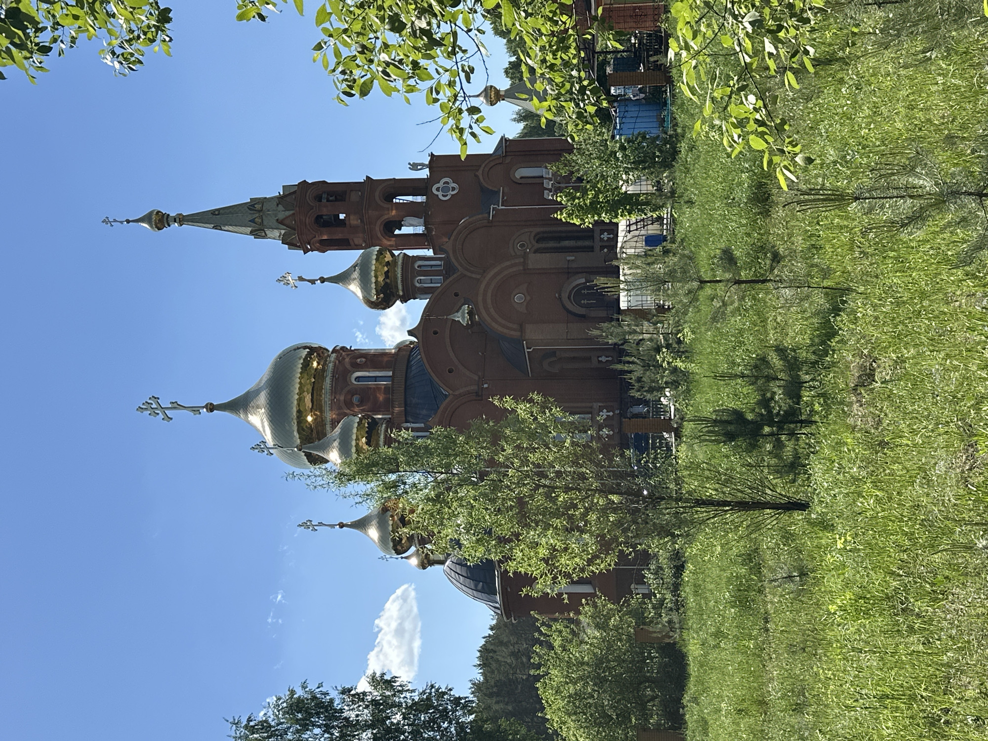 The image shows the Church of the Holy Martyr Grand Duchess Elizabeth in Chaikovsky, Russia. It is a clear day with a blue sky. The church is made of red brick and features several golden domes, some with crosses atop them. Green grass and trees are in the foreground, partially obscuring the view of the church. Leaves from trees also frame the top of the image. A building is visible behind the church.