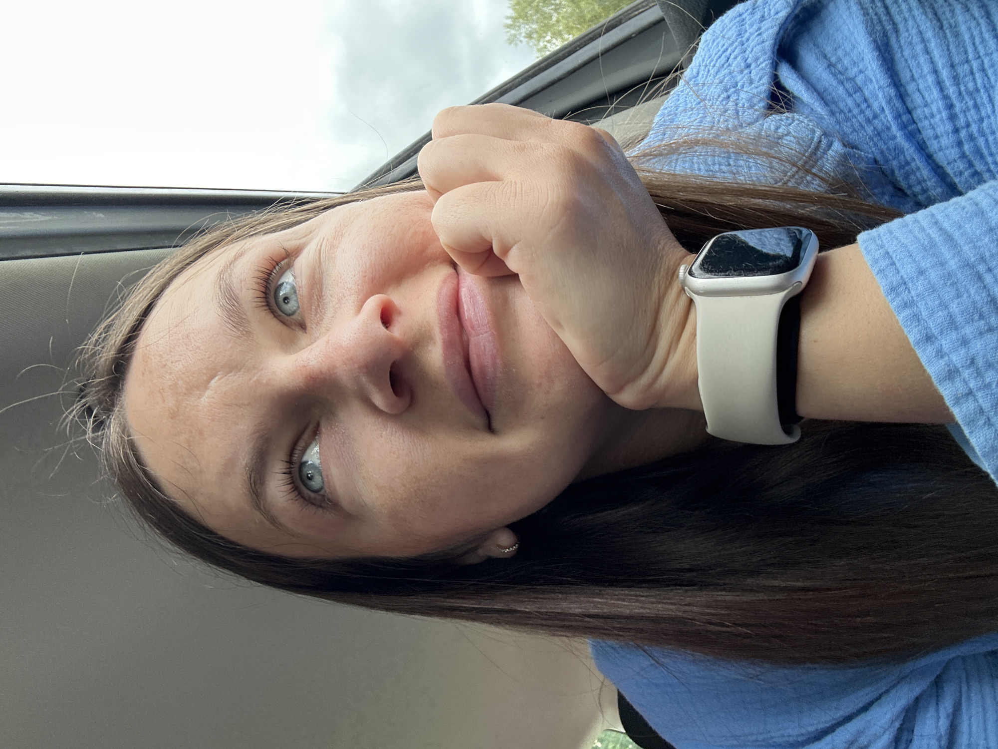 A woman with light blue eyes and brown hair, wearing a light blue textured shirt and a white Apple Watch with a black band, is posing for a selfie inside a car. She is resting her face on her hand, and the background shows a cloudy sky and the top of a tree visible through the car window. The location is unknown, as this cannot be determined from this image.
