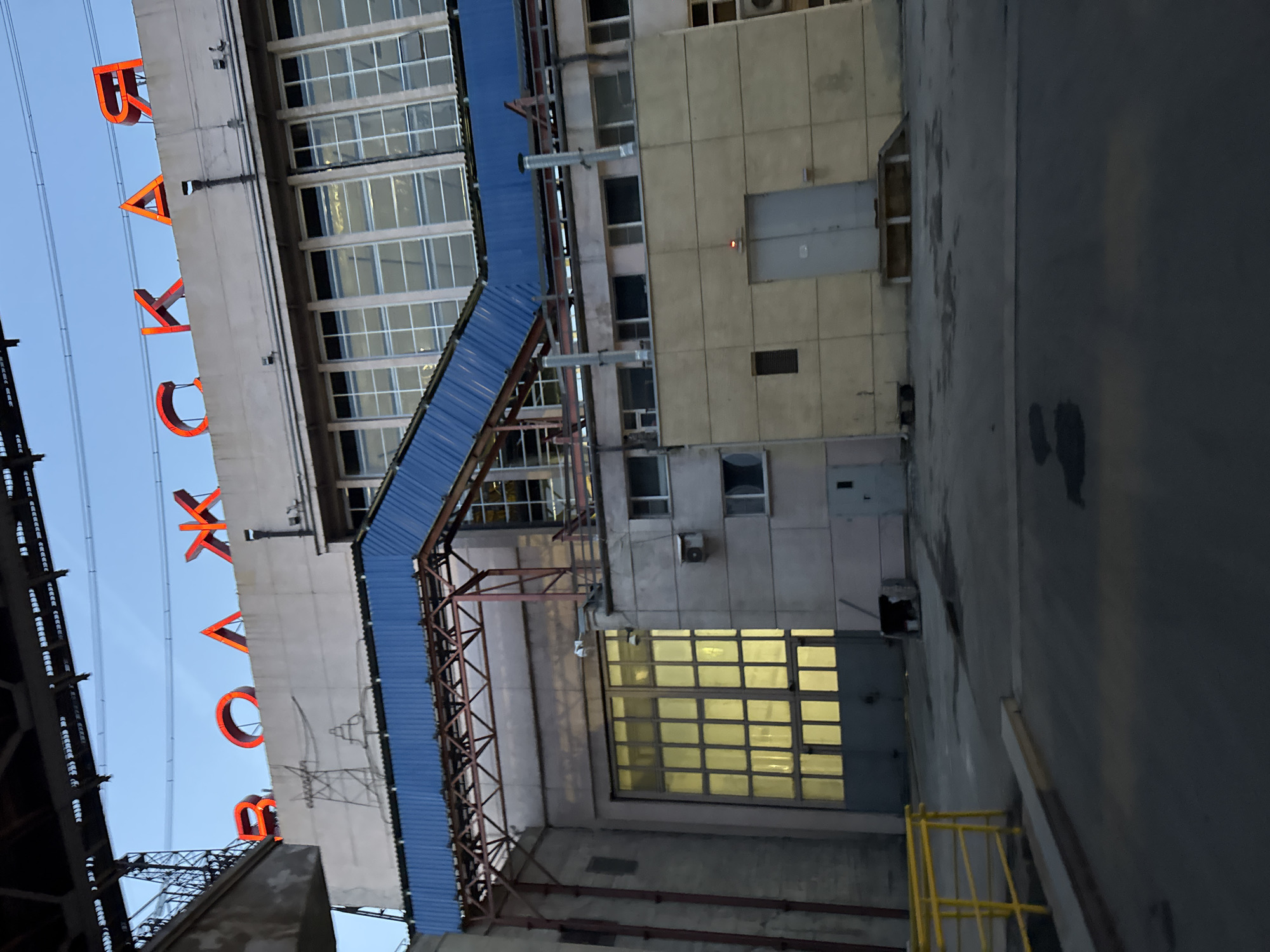 This image captures an industrial or commercial building at dusk or early night in what appears to be an urban or industrial setting in Volzhsky, Russia.
The primary subject is a multi-story building, constructed with light-colored panels and featuring numerous windows. A prominent, large, illuminated red sign spells out "БАКАХОВ" (BAKHOV or BAKAKHOV) in Cyrillic letters across the upper part of the building. The sign's glow suggests it is active, and the lighting within some of the building's windows, particularly a large ground-level window emitting a bright yellow light, reinforces the time of day as evening.
A blue corrugated metal structure, possibly a canopy or enclosed walkway, extends diagonally from the building, supported by a brown metal truss framework. Various utilitarian details such as pipes, vents, and electrical boxes are visible on the building's facade. The sky to the left of the building is a clear, light blue, indicating fair weather. To the right, a dark, paved area, possibly a road or loading dock, is visible, along with a bright yellow metal railing in the lower right corner. No people or specific activities are visible in the scene, which has a functional, somewhat stark aesthetic.