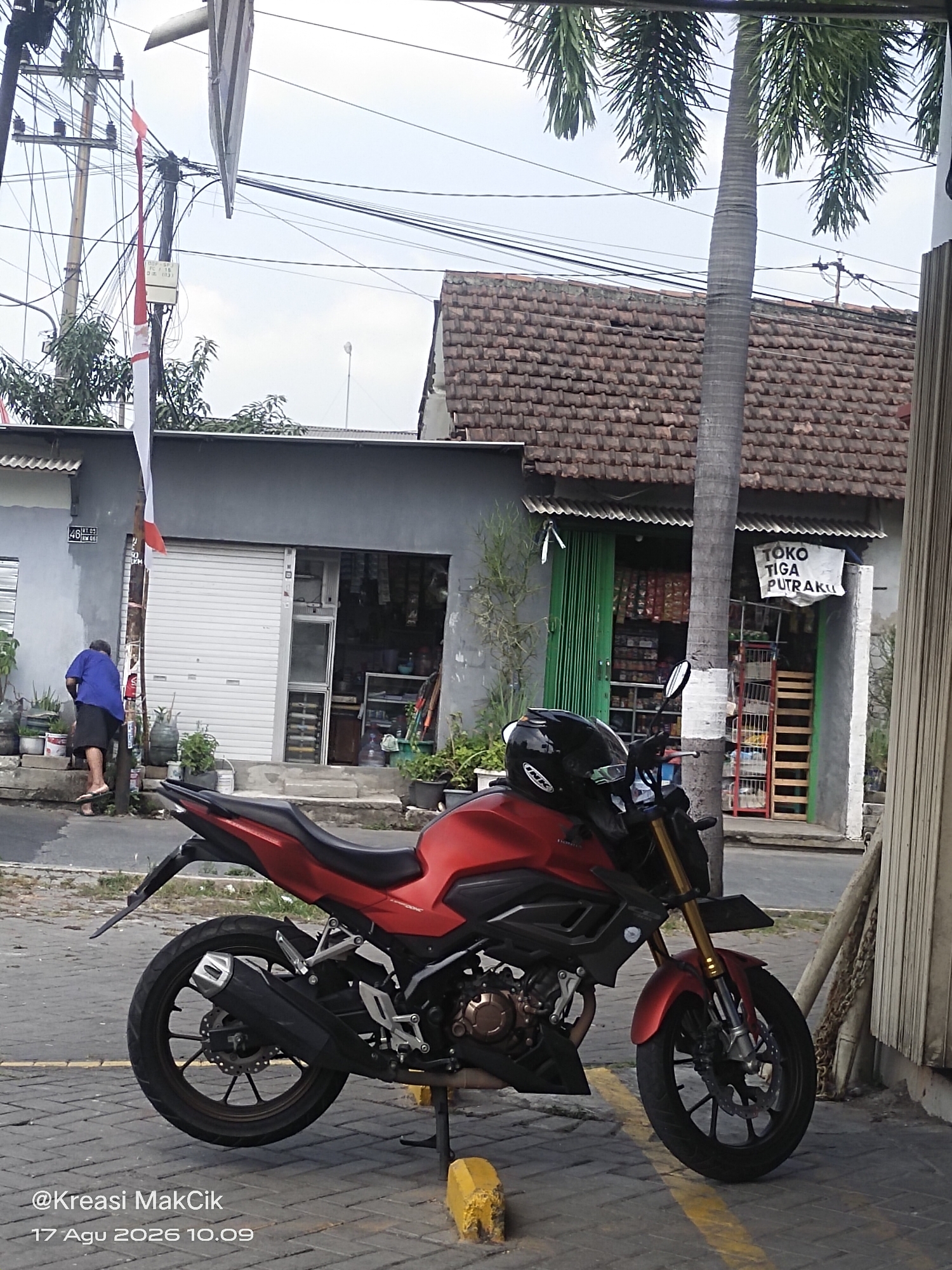 A red motorcycle is parked on a cobblestone street in front of a small shop. The shop has a tiled roof and a green corrugated metal door. A sign above the door reads "TOKO TIGA PUTRAKU." A person wearing a blue shirt and shorts is walking up a set of stairs to the left of the shop. There are several potted plants and a utility pole with electrical wires in the foreground. The sky is overcast. The timestamp in the bottom left corner indicates the image was taken on August 17, 2026, at 10:09 AM.