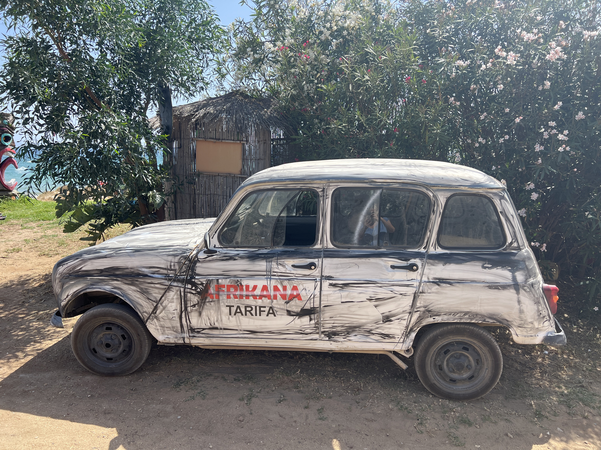 An old, white Renault 4 with a distressed finish, decorated with abstract black paint strokes and red lettering that reads "AFRIKANA TARIFA", is parked on a dirt road. The car is positioned in the foreground, facing right, with its doors and windows visible. A person can be seen reflected in the driver's side window. Behind the car, there is a rustic structure with a thatched roof and bamboo walls, suggesting a beachside or rural setting. Lush green trees and flowering bushes surround the scene, with a glimpse of the blue sea and sky in the background. The overall impression is that of a quirky, possibly vintage, establishment in or near Tarifa, Spain, a town known for its windy beaches and water sports.
