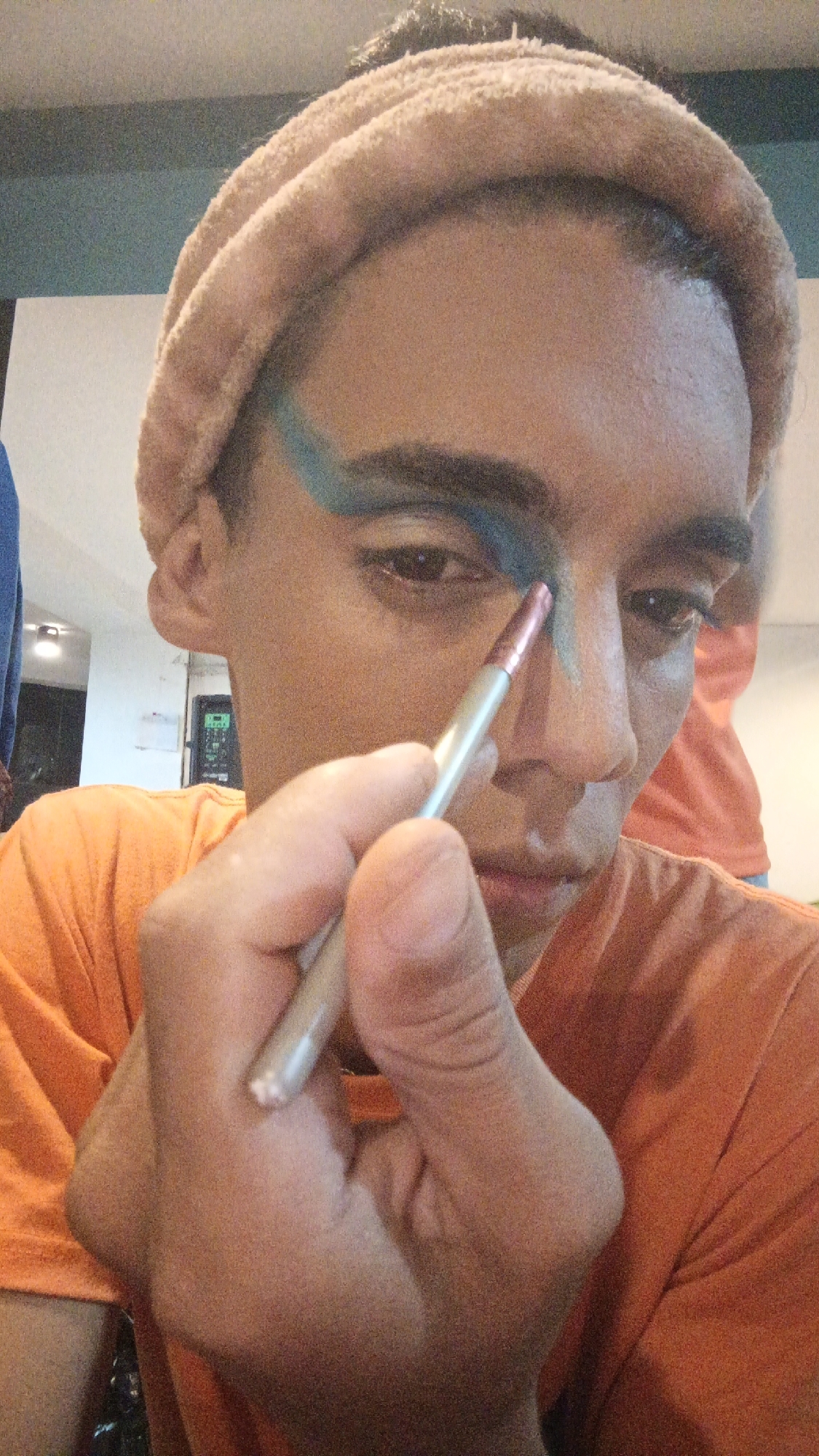 A person wearing an orange t-shirt and a tan-colored headband applies blue and green makeup to their left eyebrow with a makeup brush. The person looks down. The location is likely indoors. The city and country are only provided in the prompt, not evident in the image.