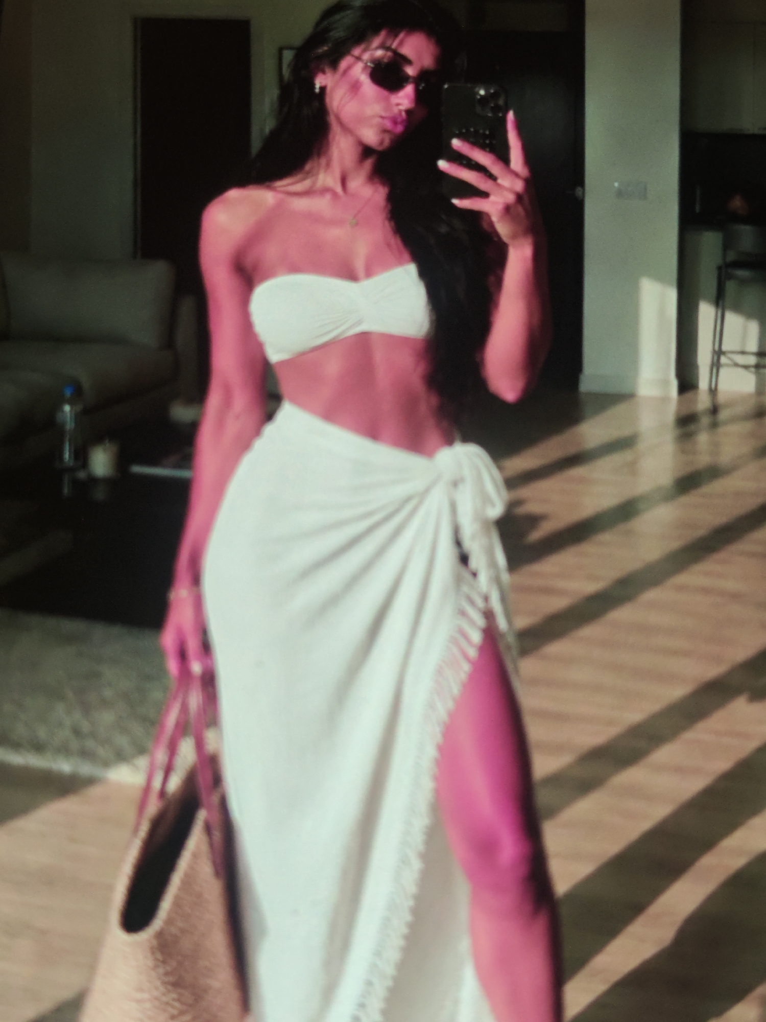 A woman with long, dark hair and sunglasses is taking a mirror selfie indoors. She is wearing a white strapless bikini top and a white sarong with a high slit. She is holding a woven tote bag in her left hand. The background appears to be a living room with a couch and a table. Sunlight is casting diagonal shadows across the wooden floor. The location is unknown.