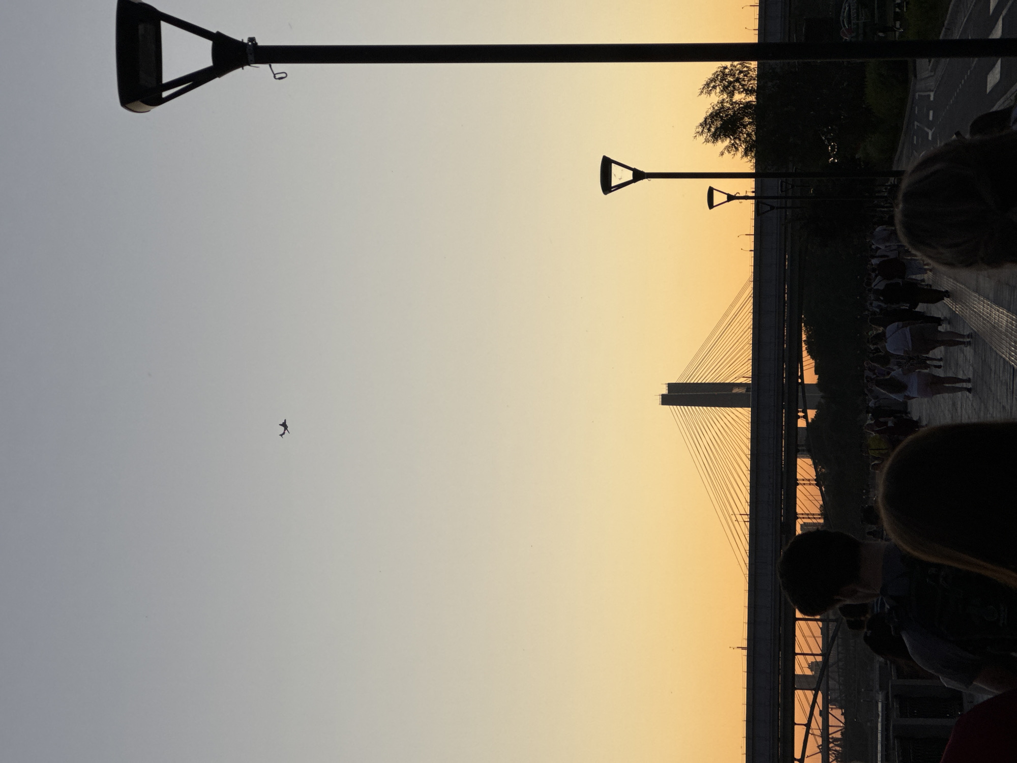 The image depicts a sunset scene in Novosibirsk, Russia. A pedestrian walkway is visible in the foreground, with a crowd of people walking along it. In the background, a bridge spans across a body of water, and a silhouetted airplane is visible in the sky. The sky is a gradient of orange and grey, indicating the time of day is sunset. The overall atmosphere is peaceful and serene.