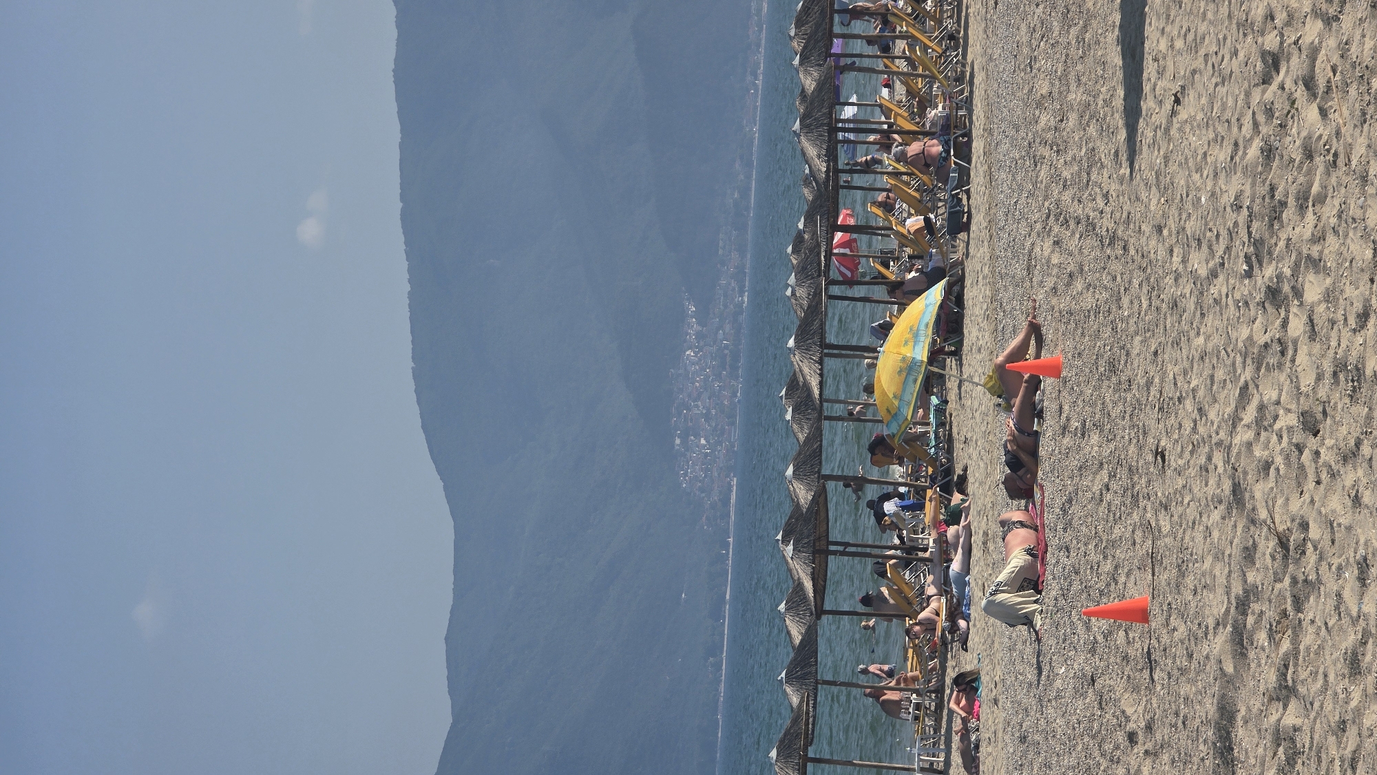 The image captures a sunny daytime scene on a sandy and pebbly beach in Asprovalta, Greece. The sky is a light, clear blue with a few small, faint white clouds. In the background, a range of dark blue-green mountains spans the horizon across a calm body of water, which is a deep blue hue. A settlement or town with numerous buildings is visible on the lower slopes of these distant mountains.
The sea contains several individuals swimming and standing in the shallows. Along the water's edge, a continuous row of straw-thatched beach umbrellas provides shade for an array of yellow sun loungers, many of which are occupied by people relaxing.
The sandy and pebbly beach occupies the foreground and mid-ground. Several individuals are sunbathing directly on the sand. One person is lying on a red mat under a yellow and blue striped beach umbrella. Another person lies on the sand with a light-colored fabric covering their lower body. Two orange traffic cones are positioned in the sand in the foreground; one is closer to the viewer, and the second is placed beside a person sunbathing.