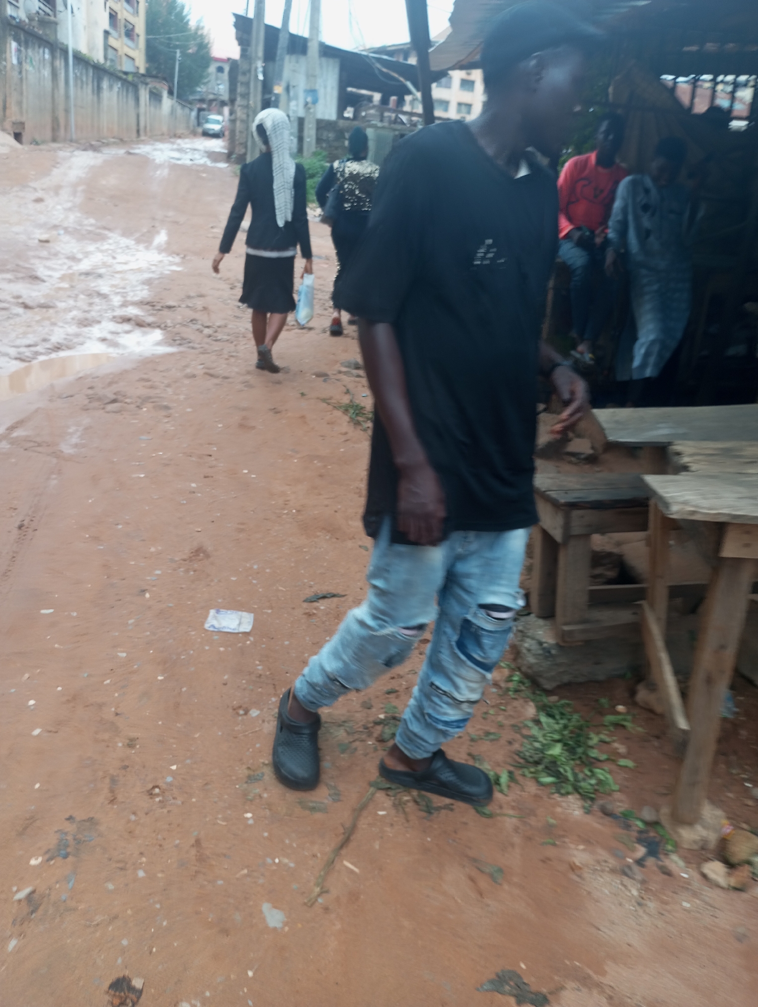 In Onitsha, Nigeria, a dusty, unpaved road with puddles of water suggests recent rain. A person wearing ripped light blue jeans and black Crocs walks toward the viewer. They have a dark complexion, wear a black t-shirt and a dark baseball cap. Behind them, two other individuals are visible: one in a black jacket and skirt, wearing a white headscarf, and another wearing a dark jacket with gold embellishments. Further in the background, three men are seated in a shaded area with wooden tables, one man wearing a bright orange shirt. The environment is urban, with buildings and utility poles lining the street.