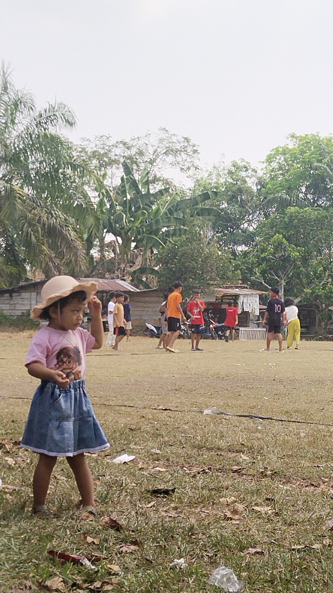 In the city of Margatunggal, Indonesia, a young girl in a light-colored sun hat, pink t-shirt with a graphic print, and a blue denim skirt stands in a grassy field. She is looking to her left and appears to be touching her hat. In the background, a group of children and a few adults are gathered on the field. Some children are wearing t-shirts and shorts, and one is wearing a black shirt with the number 10 on the back. There are trees and some simple structures with corrugated metal roofs in the background. The ground is dry and covered with patches of grass, fallen leaves, and some scattered white plastic debris. Two motorcycles are parked to the right of the background structures. The sky is overcast.