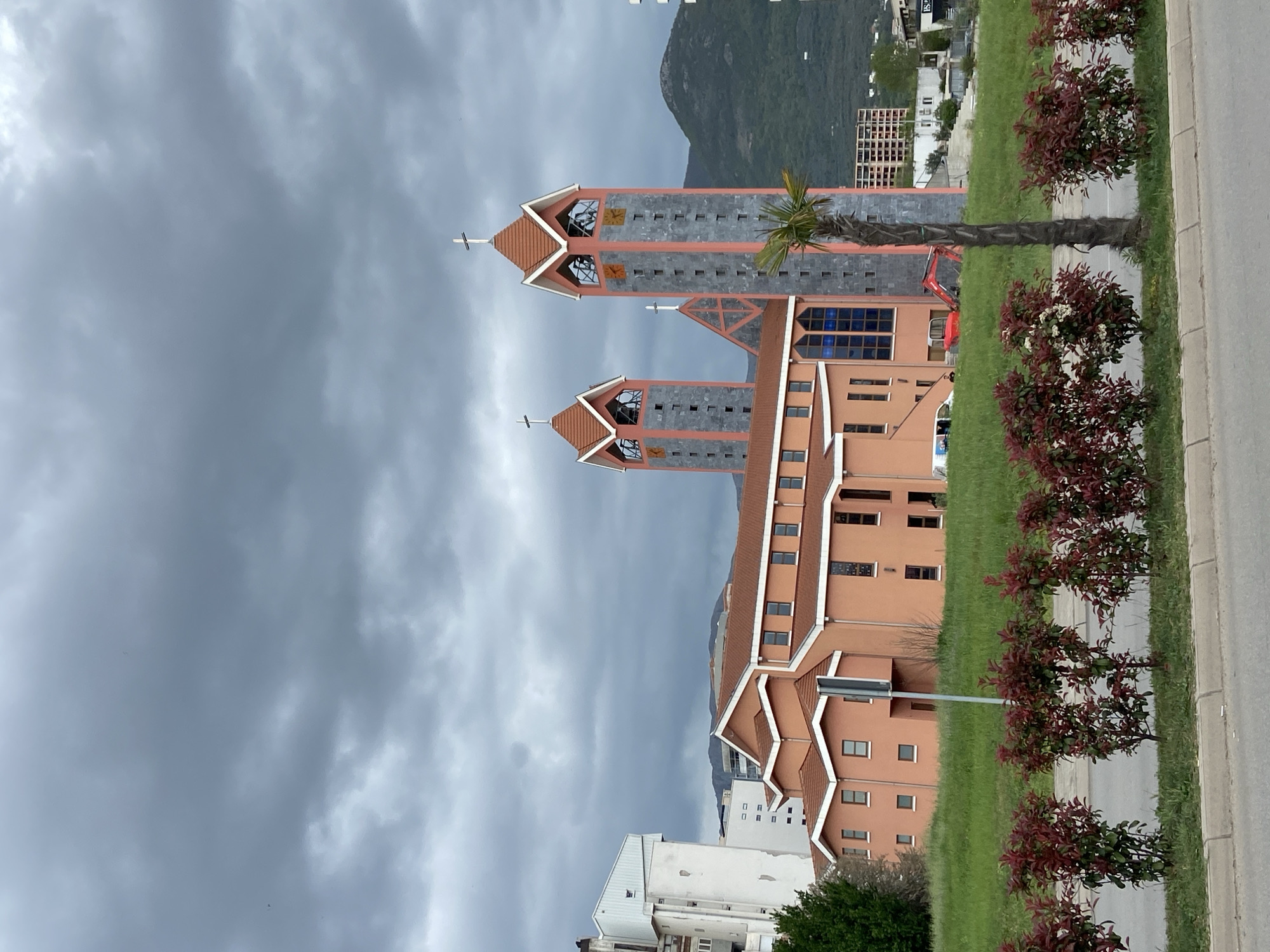 The image shows a modern, salmon-colored church with two prominent towers located in Bar, Montenegro. The church towers are gray with orange trim and topped with peaked roofs and crosses. The main body of the church has a similar color scheme with visible windows and architectural details. A palm tree and other greenery are positioned beside the church. In the background, a mountain is visible under a cloudy, overcast sky. A road and sidewalk are also in the foreground with trimmed bushes along the edge. Adjacent buildings are visible, indicating an urban environment. A piece of construction equipment can be spotted near the base of the tree.