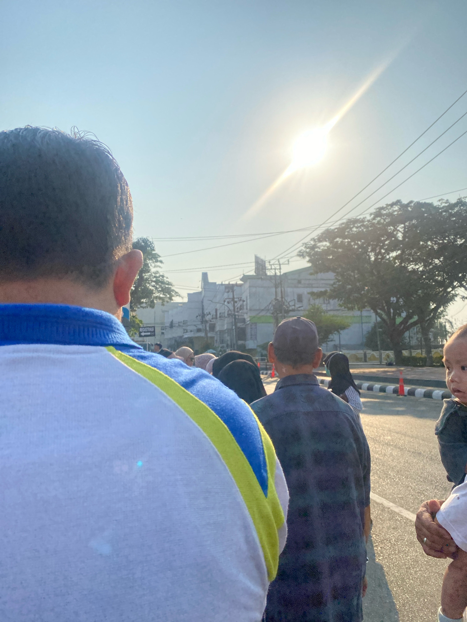 The image shows a group of people standing on a street in what appears to be a city. The sun is shining brightly, creating lens flare. In the foreground, a person wearing a blue and white shirt with yellow trim is seen from behind. Further back, there are several other individuals, some wearing headscarves, and a child. A building with windows and signs, including one that says "REGISTRASI," is visible in the background, along with trees and utility poles. The street has a marked lane and a striped curb. The overall scene suggests an outdoor gathering or event in an urban environment. The "REGISTRASI" sign, meaning "registration" in Indonesian, and the presence of people in modest attire, potentially attending a religious or community event, are indicators. This scene is consistent with a gathering in Banjarmasin, Indonesia, possibly related to a local festival or public event.
