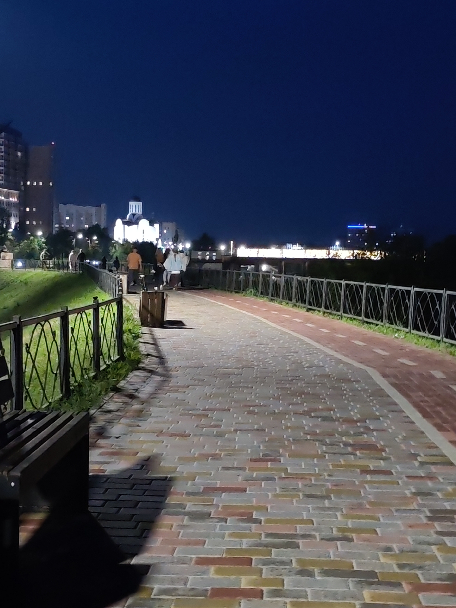 In this nighttime scene in an urban park, a brick pathway with a red border curves to the right, leading away from the viewer. The pathway is illuminated by streetlights and the lights of distant buildings, creating shadows from a bench on the left and a trash bin. The pathway is bordered by a decorative metal fence on the left and a grass embankment with a fence on the right. Several figures are visible walking and standing on the pathway and embankment. In the background, city buildings, including a white domed structure resembling a church and a high-rise building with lit windows, are illuminated against a dark blue sky. The environment suggests a public space designed for evening strolls and recreation.