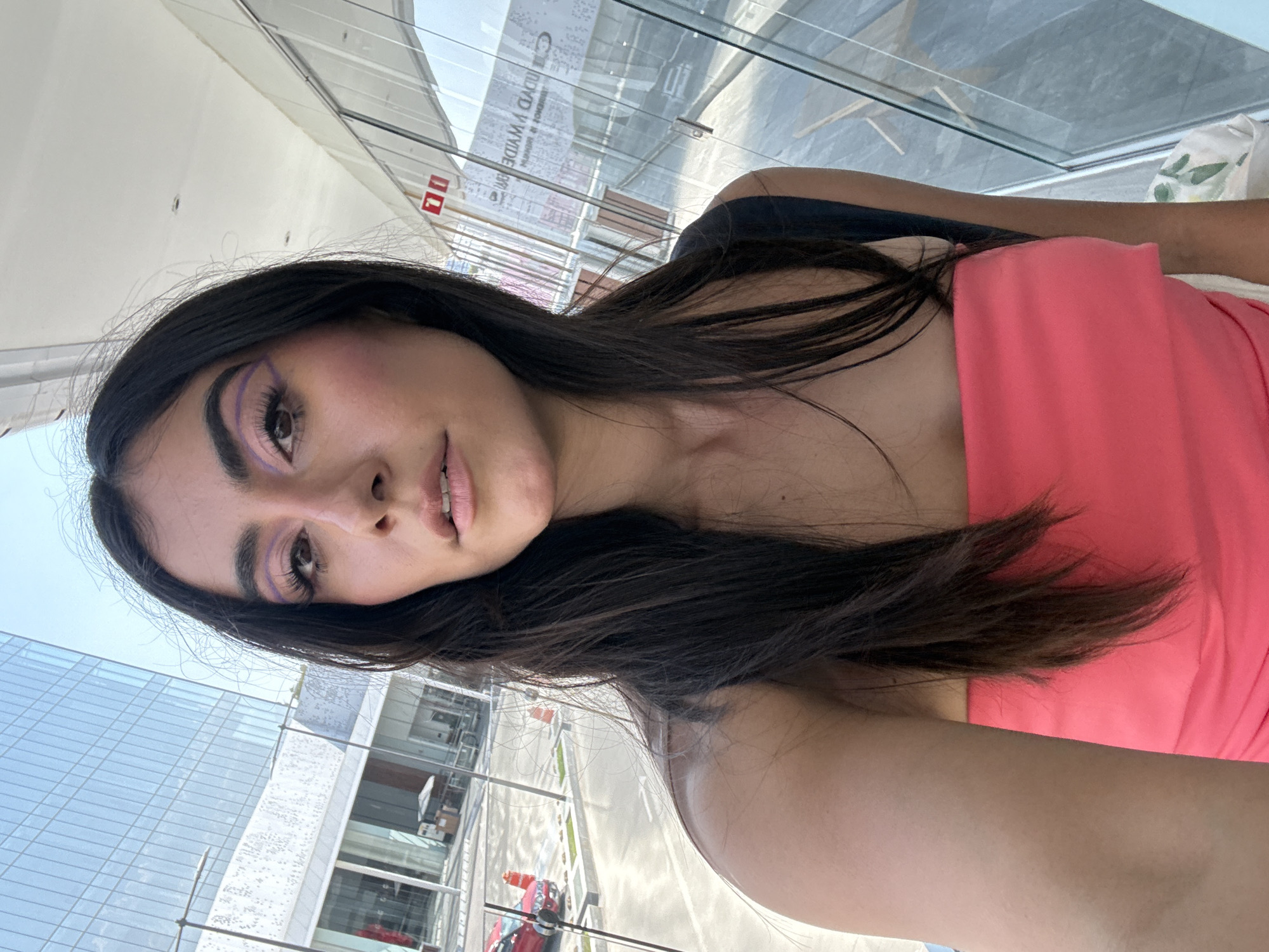 A young woman is depicted in a selfie, indoors with a large window displaying an external cityscape. The woman has long, dark brown hair and is wearing a bright pink top. She is facing the camera with a slight angle. Her makeup includes a prominent pink eyeshadow and liner.
Through the window, a modern building with a glass facade is visible, indicative of an urban setting. Additionally, streets and what appear to be vehicles are visible in the exterior view. Based on the architecture and context clues, the location is identified as being in the city of Puebla, Mexico.