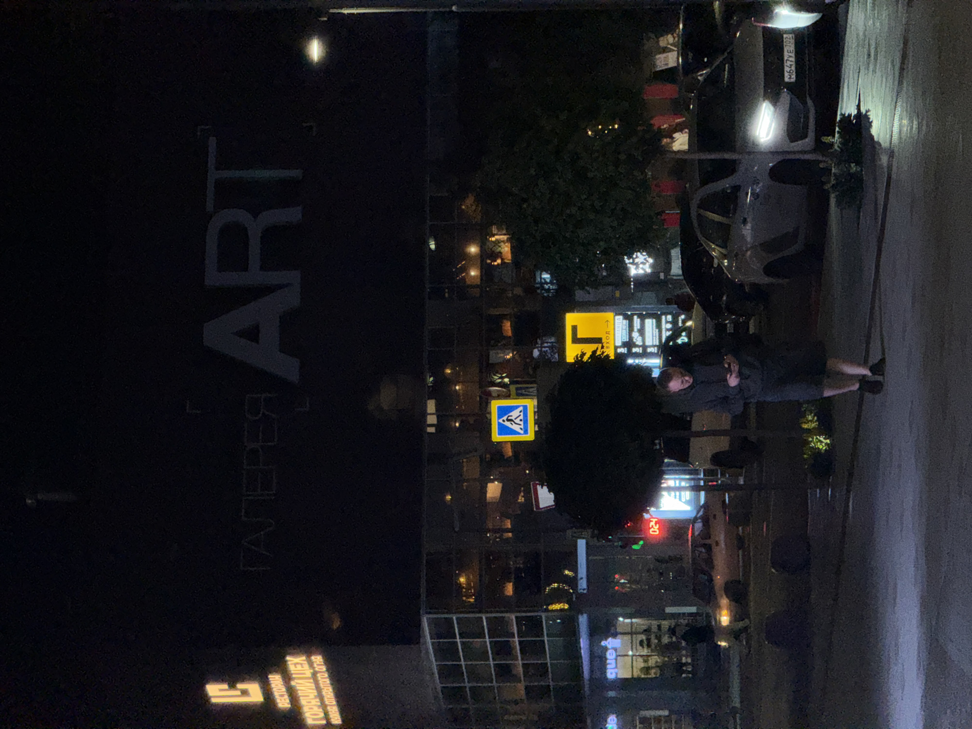 The image depicts a nighttime street scene in Ufa, Russia. In the background, a large building is illuminated, featuring signage that reads "ГАЛЕРЕЯ ART" (Gallery ART) and "РЕСТОРАН ГОРЯЧИЙ ЦЕХ КУХНЯ ОТКРЫТОГО ОГНЯ" (Restaurant Hot Workshop Open Fire Kitchen). A pedestrian crossing sign is visible. In the foreground, a man is walking while looking at his phone. Several cars are parked on the street. The overall environment is urban, with streetlights and building lights providing illumination.