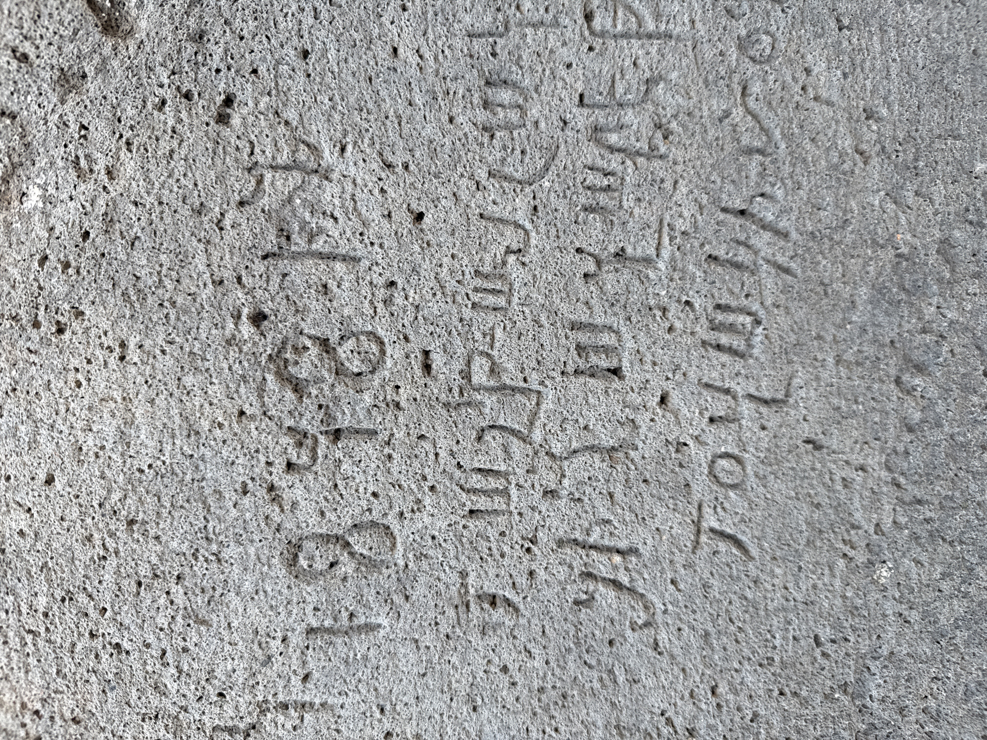The image displays a close-up view of an inscription carved into a rough, porous, grey stone surface. The inscription appears to be in Armenian script, with some characters clearly visible and others partially obscured or worn. The first line of the inscription consists of numbers: "1848". Below this, there are two more lines of Armenian script, the first line containing characters that appear to read "ԱՂԱՆԴ", and the second line containing characters that appear to read "ՀԱՅԿԱԶՆ". The stone itself exhibits a granular texture with numerous small pits and imperfections, indicative of its weathered state. The lighting suggests the inscription is outdoors, with shadows cast by the carved characters. The specific location of this inscription is at the St. Bartholomew Monastery in the village of Goght, Armenia.