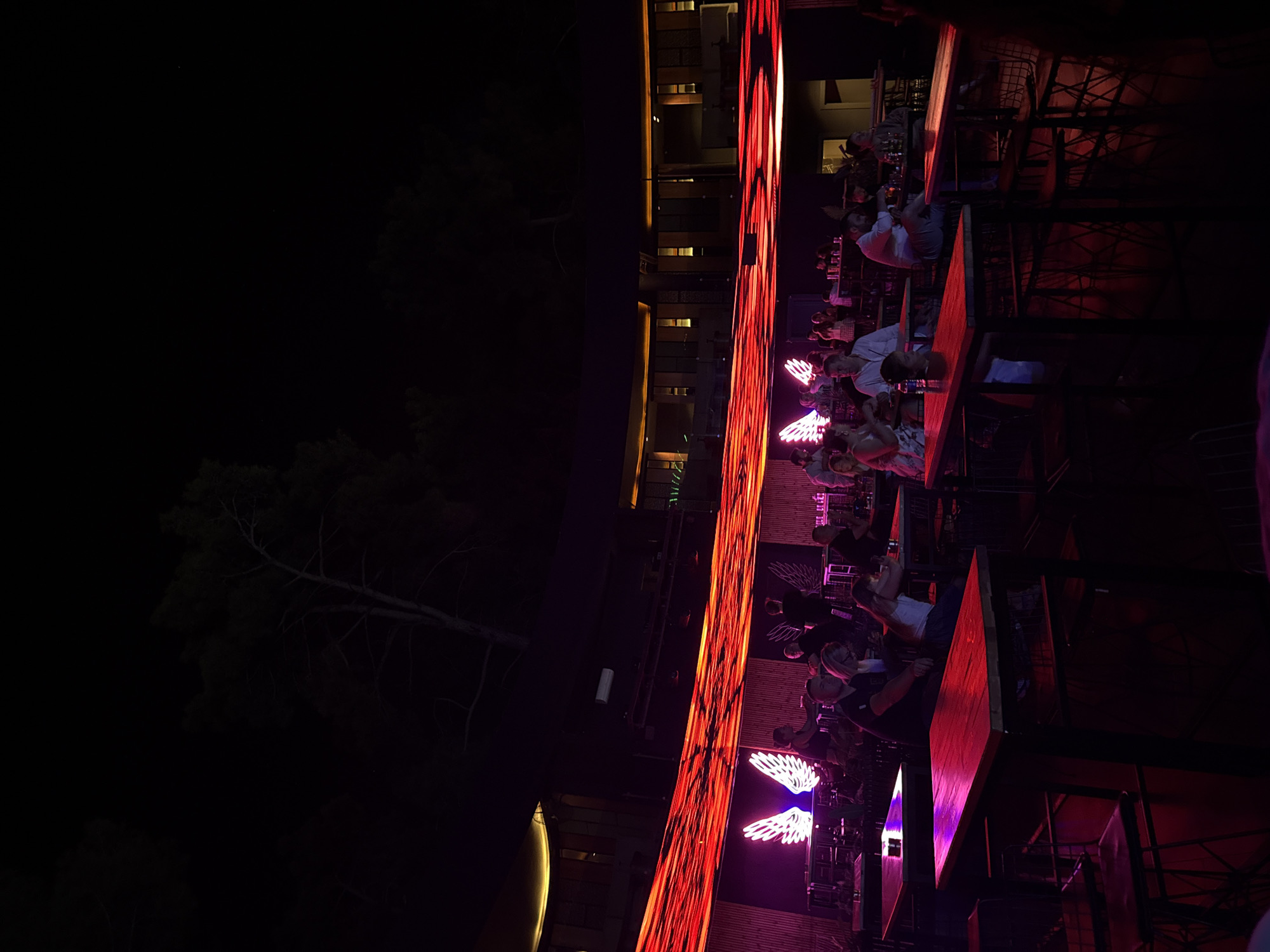 This is a nighttime photograph of a crowded outdoor seating area at what appears to be a club or restaurant in Bodrum, Turkey. The scene is illuminated by vibrant red and purple lighting, with a prominent neon sign depicting white angel wings. Patrons are seated at various tables, some engaging in conversation and others observing a performance or light show. The architecture suggests a multi-level structure with a curved facade. Trees are visible in the background against the dark night sky. The overall atmosphere is lively and suggests entertainment.
