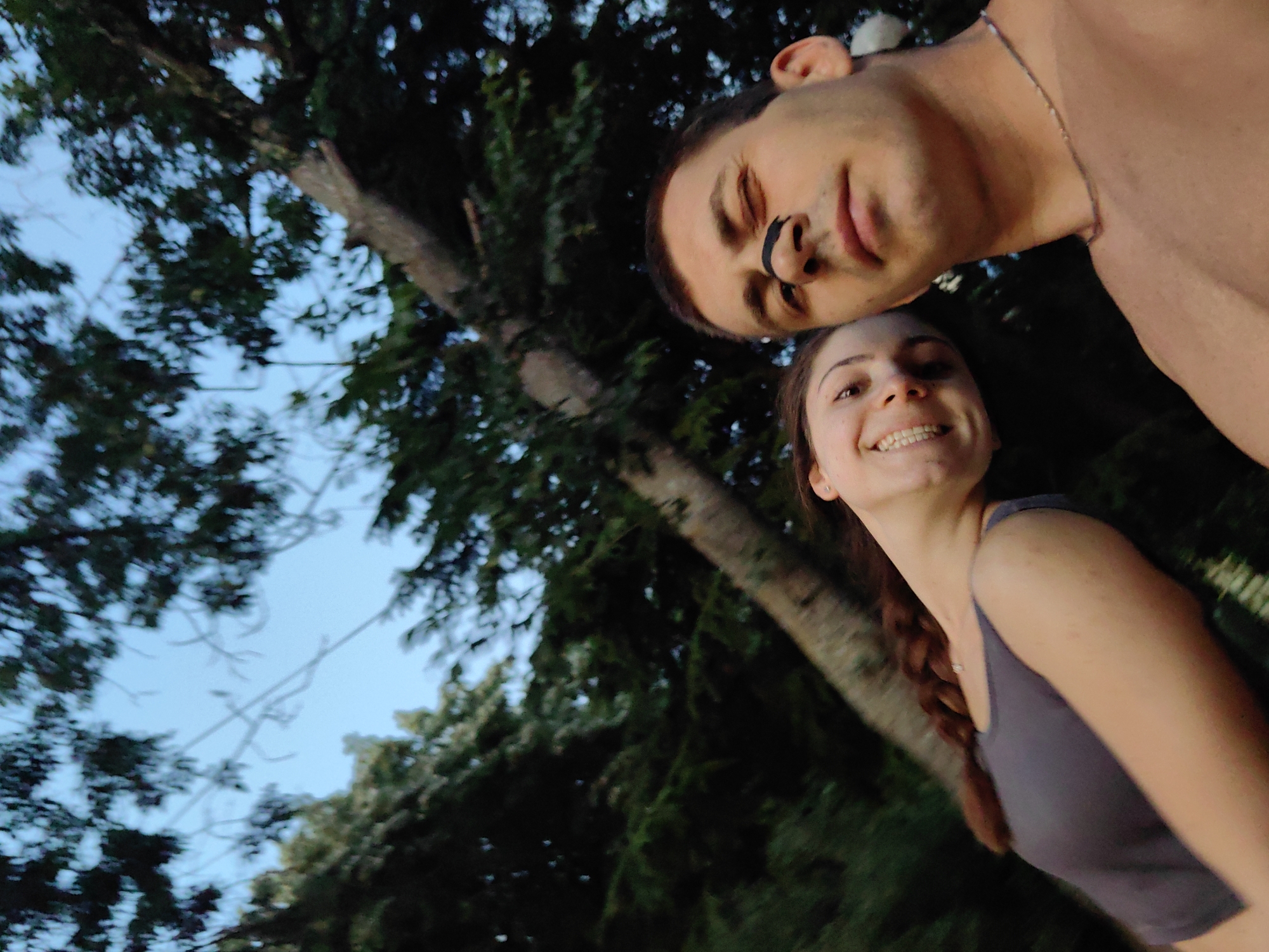 Two individuals, a male and a female, are captured in a low-angle, outdoor photograph, likely taken during daylight hours with a clear sky. The male, positioned in the upper portion of the frame, has a portion of his face visible, with his left eye closed and his nose adorned with a dark mark. He appears to be shirtless, wearing a delicate chain around his neck. The female, situated below the male, has her face turned towards the camera, smiling with her teeth exposed. She is wearing a grey tank top, and her hair is styled in a braid. The background features lush green trees, suggesting a natural setting. The image may have been taken in the vicinity of the Varna City Sea Garden, a prominent park in Varna, Bulgaria, known for its abundant greenery and proximity to the coast.