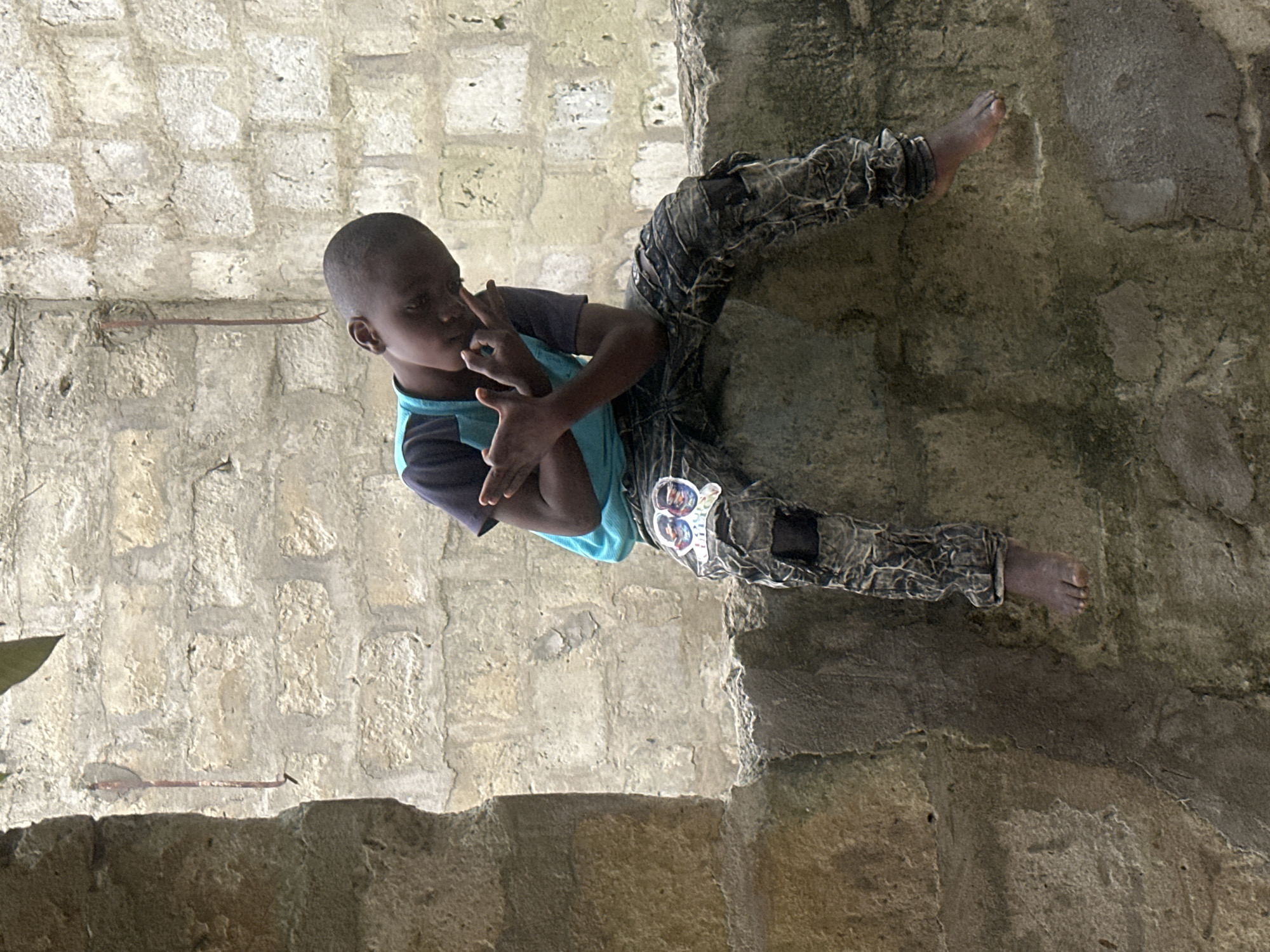 A young boy, seemingly of African descent, is depicted in a photograph taken in Badagry, Nigeria. He is positioned in a seated or resting posture, leaning against a rough, textured wall, possibly made of stone or concrete. The boy is wearing a blue t-shirt with dark accents and patterned trousers that appear somewhat worn or ripped. His gaze is directed slightly away from the camera, and his hands are near his face, with one finger touching his cheek.
The background consists of a stone-block wall, suggesting an outdoor or semi-outdoor environment. The lighting appears natural, casting shadows and highlighting the textures of the surfaces. The overall impression is one of candidness, capturing a moment in the boy's day. The context of Badagry, Nigeria, places the scene within a specific geographical and cultural setting. There is no discernible text within the image.