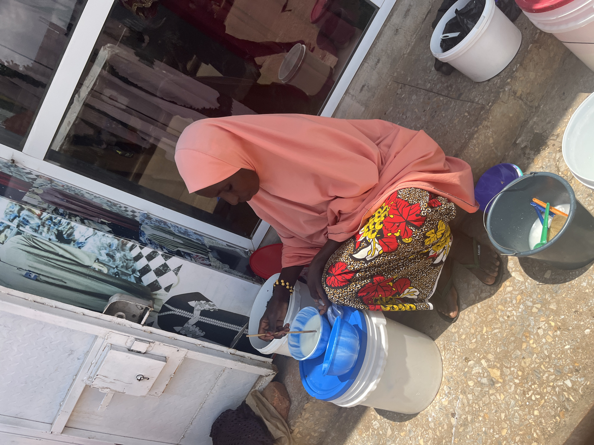 In the city of Jos, Nigeria, a young woman wearing a pink hijab and a brightly patterned skirt sits on the ground next to a shop. She is mixing a white liquid in a blue bowl with a wooden stick, which rests on a larger blue lid. In front of her are several buckets: a white one, a gray one containing a green and orange object, and another white one with a black plastic bag inside. The shop window behind her displays clothing and various mannequins. The ground is a rough, speckled concrete.