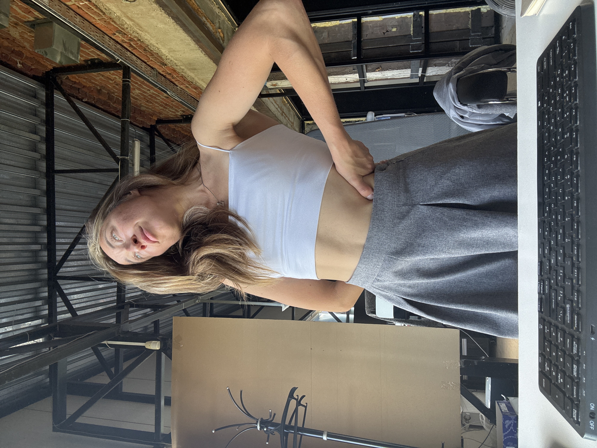 A young woman with long, blonde hair is standing in what appears to be a studio or workshop space. She is wearing a white crop top and a grey pleated skirt. Her right hand is on her hip, and she is looking upwards and to the left with a pensive expression. The background features exposed brick walls, a corrugated metal ceiling with exposed beams and ductwork, and what looks like a coat rack. A desk with a keyboard is visible in the foreground. The image appears to be taken in Chaikovsky, Russia, given the context provided.