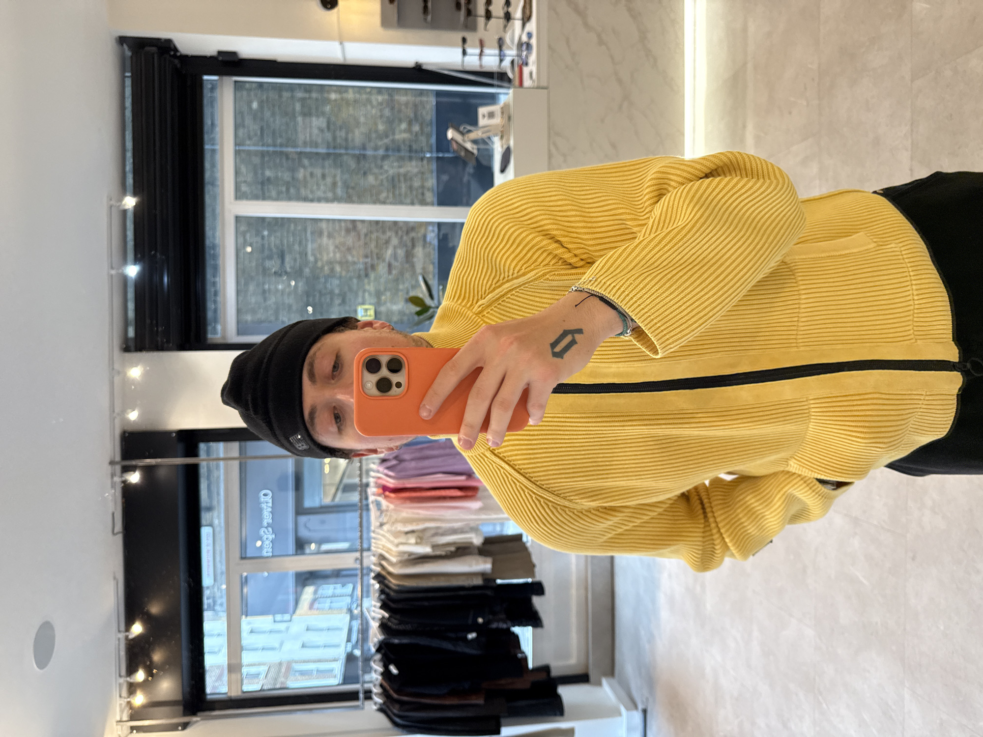A young man takes a mirror selfie inside what appears to be a clothing store. The image is oriented vertically, with the man's head on the left and his body extending to the right. He wears a vibrant yellow ribbed long-sleeved top with a black central zipper and a black beanie. He holds an orange smartphone in his right hand, positioned to capture his reflection. Visible on his left forearm is a black tattoo resembling a stylized 'D' or 'G', and he has multiple thin bracelets on his wrist.
The background of the reflection reveals the interior of the store, featuring clothing racks with neatly folded garments in various colors such as purple, pink, white, beige, and black. A large window on the left side of the reflection shows the exterior, including buildings and a street scene. Mirrored text "Oliver Spencer" is clearly visible on the window, indicating the brand or store name. The lighting suggests it is daytime, with overhead lights illuminating the store interior. The floor is light-colored and reflective, contributing to the bright atmosphere. The overall setting implies a retail environment, consistent with the provided context of London, United Kingdom, where Oliver Spencer is a known brand.