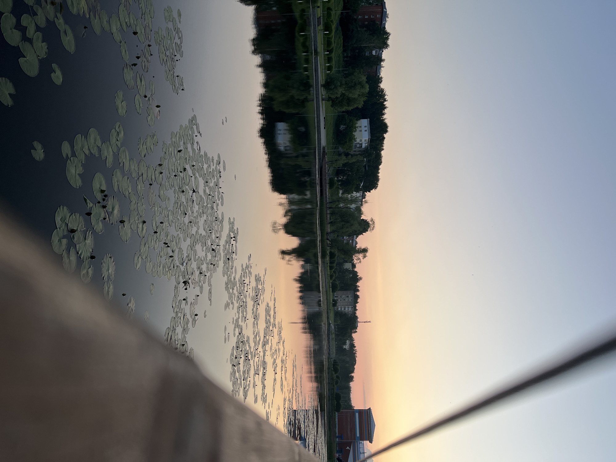 The image depicts a serene waterfront scene in Votkinsk, Russia, during twilight. A calm body of water, likely a lake or river, dominates the foreground, its surface dotted with numerous water lily pads. The water reflects the soft hues of the sunset sky, creating a gentle gradient of pale blue to soft orange.
Across the water, a tree-lined embankment is visible, with residential buildings of varying heights nestled amongst the greenery. The architecture suggests a blend of Soviet-era apartment blocks and perhaps some newer constructions. Further along the embankment, a modern building with red brick and blue accents stands out, possibly a public facility or sports complex. A telecommunications tower is also visible in the distance.
The perspective is from a slightly elevated vantage point, possibly a bridge or boardwalk, with a blurred wooden railing or structure in the immediate foreground on the right side of the frame. The sky is clear, with the last vestiges of sunlight painting the horizon. No people are visible in the scene, contributing to the tranquil and undisturbed atmosphere. The overall impression is one of peaceful urban nature at the close of day.