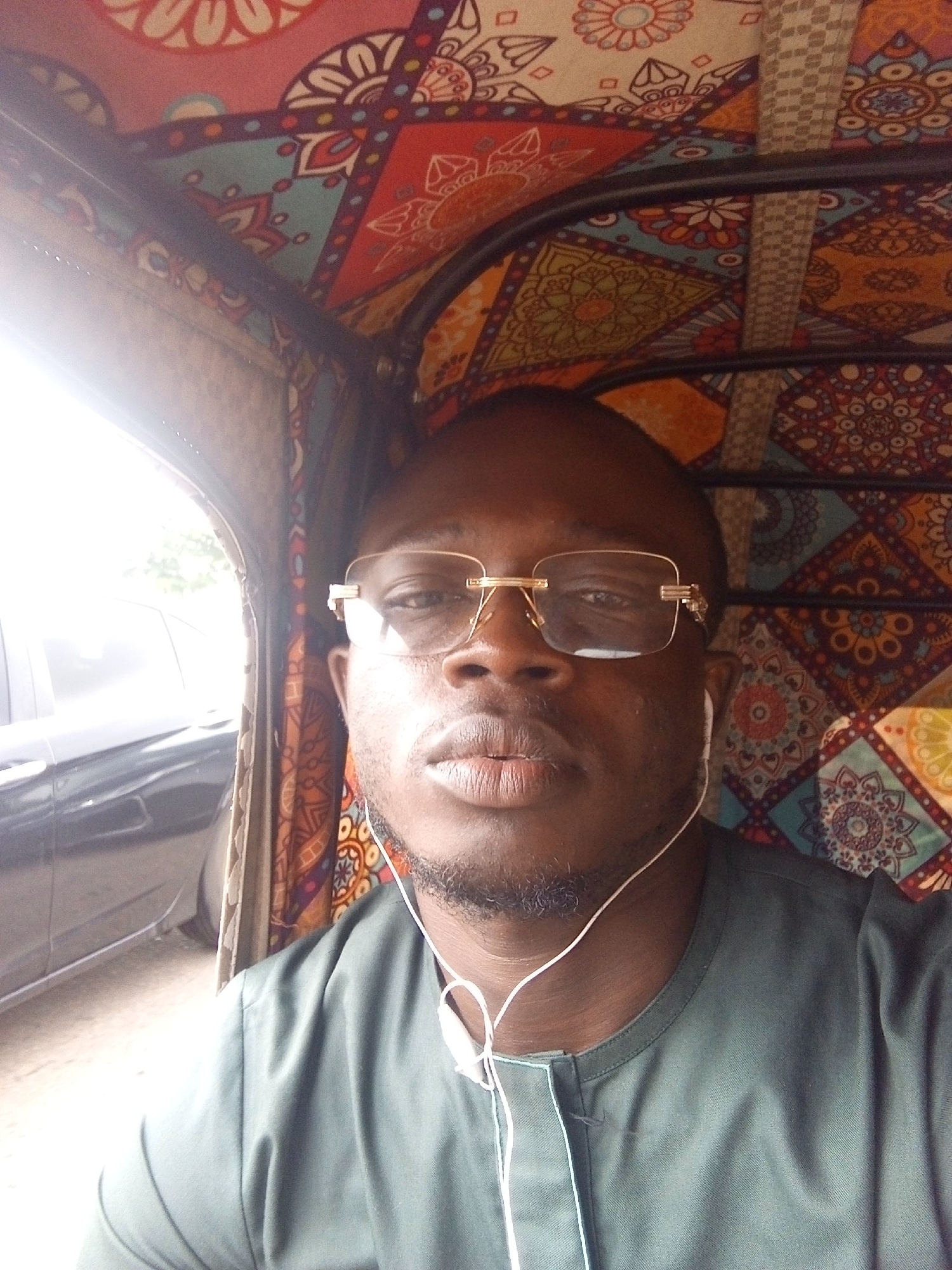 A dark-skinned man is captured in a medium close-up selfie, seated inside a vehicle during daytime. He looks directly at the camera with a neutral expression. He wears prominent gold-rimmed rectangular glasses and has white wired earbuds hanging around his neck and partially in his ears. His attire consists of a dark green, traditional-style top with a simple, closed placket neckline. He has a short, well-maintained beard.
The interior of the vehicle is vibrantly decorated with a patterned fabric featuring intricate geometric and floral designs in various colors including red, orange, blue, green, and white. This distinct upholstery is commonly found in auto-rickshaws (Keke Napep) in Nigeria. To the man's left, a bright outdoor scene is visible through an open side of the vehicle, showing a glimpse of a dark-colored car and suggesting a street or roadside environment. The intense light from the left indicates a sunny day. No visible text is present in the image. The setting is consistent with locations like Galadimawa Village, Nigeria.