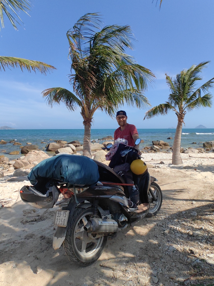 A man sits on a scooter on a sandy beach with palm trees, the ocean, and a clear blue sky in the background. The scooter is loaded with a dark blue pack and a yellow helmet hangs from the handlebars. The man wears a red shirt and a baseball cap. The license plate is visible on the back of the scooter. Rocks are scattered along the shoreline.