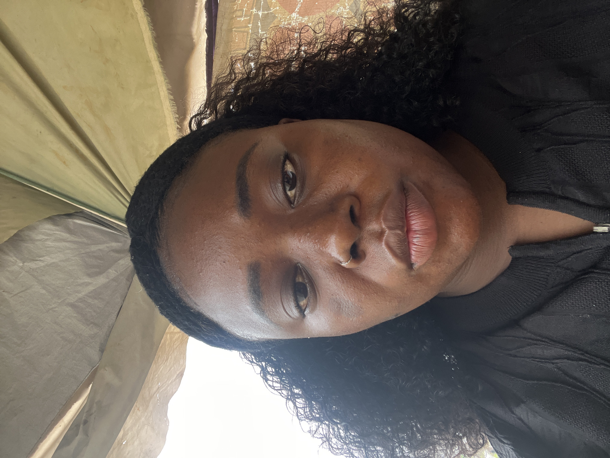 The image is a close-up, front-facing selfie of a Black woman with dark, curly hair. She is wearing a black top. Her facial features include dark eyes, a visible nose ring, and full lips. The background appears to be outdoors, possibly under a canopy or awning, with a textured, yellowish fabric overhead and some indistinct structures to the right. The lighting suggests it is daytime. The subject's expression is neutral. There is no visible indication of the location being the city of Abuja, Nigeria.