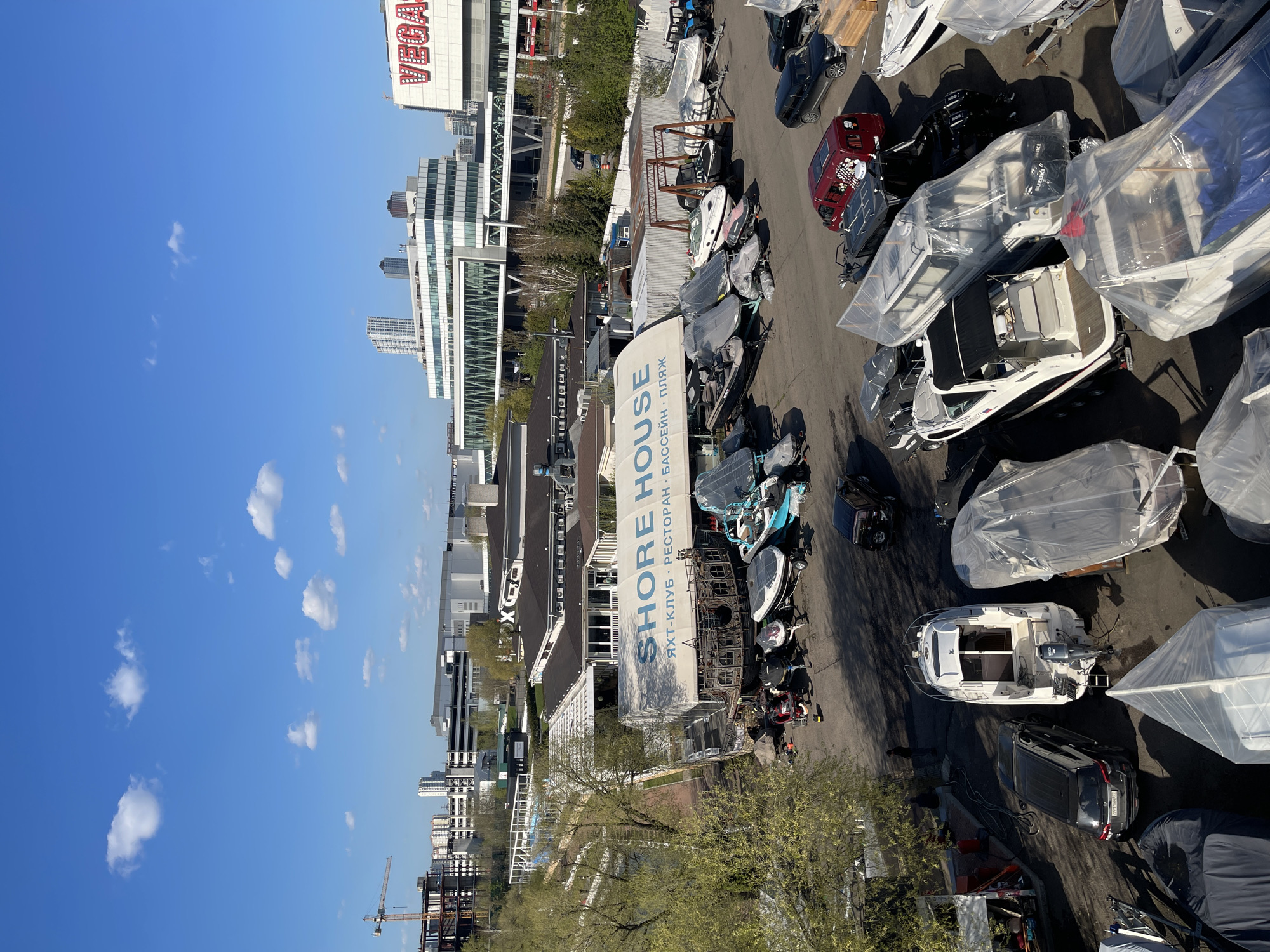The image shows an aerial view of a boat storage area located in Khimki, Russia, near the Shore House Yacht Club. Several boats, some covered in plastic, are parked on the asphalt. A building with a sign reading "Shore House" in both English and Russian is visible. Adjacent to the boat storage are residential buildings and skyscrapers, indicating a modern urban landscape. The sky is blue with scattered clouds. A construction crane is present, suggesting ongoing development in the area.