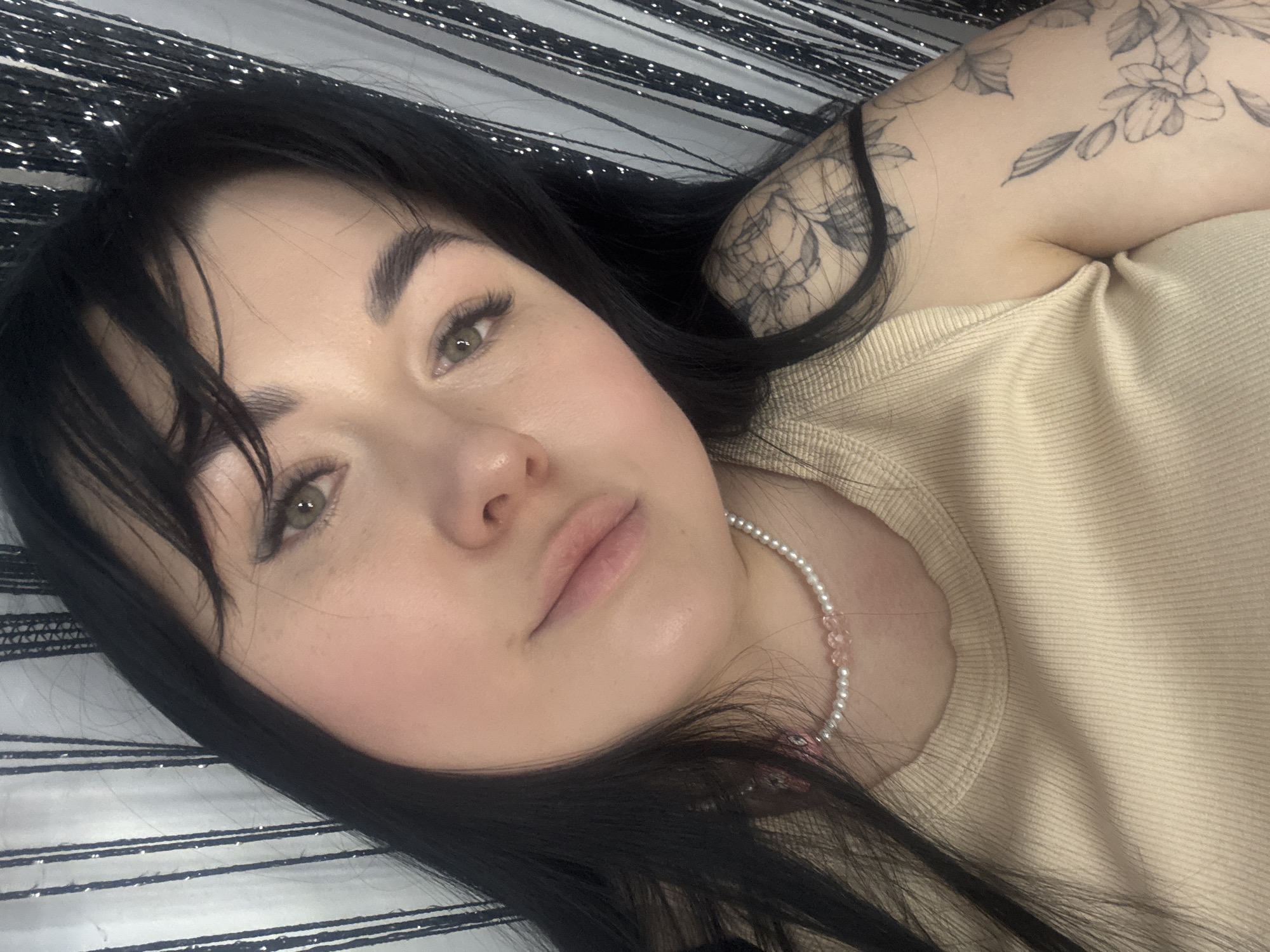 A close-up, top-down shot features a fair-skinned woman with dark, straight hair and a tattoo on her right shoulder. She is wearing a light beige ribbed shirt and a pearl and pink bead necklace. The background consists of silver and black striped material. The location is not discernible based on the image alone, and cannot be determined to be in Chaikovsky, Russia.