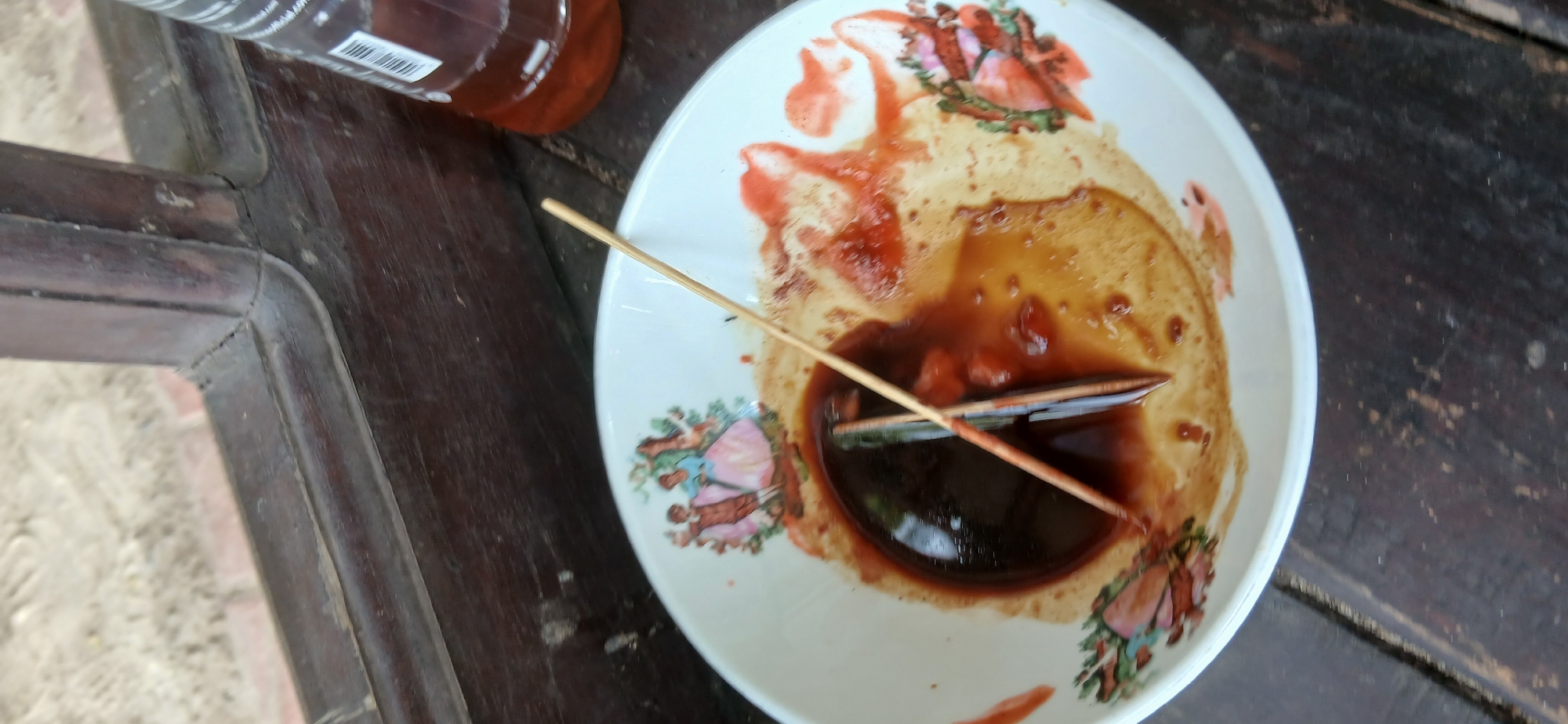 A close-up, overhead shot captures a white ceramic bowl, decorated with painted figures of people in a garden scene, resting on a dark wooden surface. Inside the bowl, a dark brown liquid, likely soy sauce or a similar condiment, pools at the bottom. Scattered around the dark liquid are streaks of red, possibly from chili sauce or a similar ingredient, mixed with lighter brown residue. Two wooden skewers are partially submerged in the liquid, one crossing over the other. To the right of the bowl, a portion of a clear plastic bottle containing a reddish-brown liquid is visible. The background shows a blurred view of a wooden window frame and sandy ground.