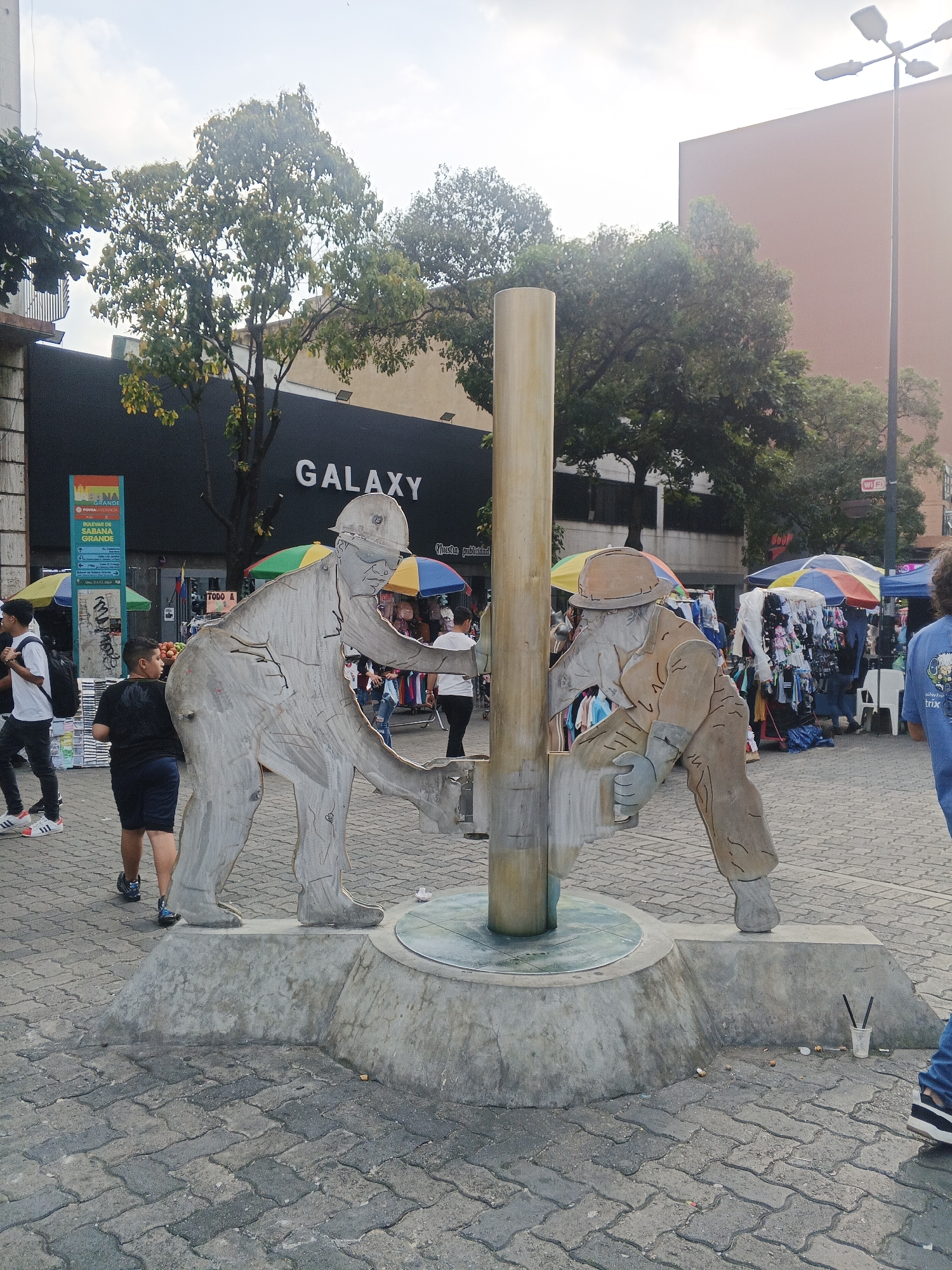 This image captures a vibrant street scene on the Bulevar de Sabana Grande in Caracas, Venezuela, during the daytime. The central focus is a unique statue featuring two workers, depicted as flat, cutout figures in hard hats, interacting with a tall, cylindrical column. The statue, made of a textured material like wood or metal, is mounted on a concrete base amidst a paved pedestrian area.
In the background, numerous people are seen walking and browsing. The area is bustling with activity, characterized by a series of market stalls shaded by colorful umbrellas, selling what appear to be clothes and other goods. Behind these stalls, a large building prominently displays the word "GALAXY" in white letters against a dark facade. Other visible text includes "BULEVAR DE SABANA GRANDE" and "PONSALADA" on a green sign, "TODO A" on a white sign, and a small "WIFI" sign on a lamppost. A person on the right wears a blue shirt with "ailwhp trix" printed on it.
The sky is bright and partly cloudy, illuminating the scene and the scattered green trees. The ground is paved with interlocking bricks. Some litter, including cigarette butts and a plastic cup with straws, is visible around the base of the statue, hinting at the public and active nature of the location.
