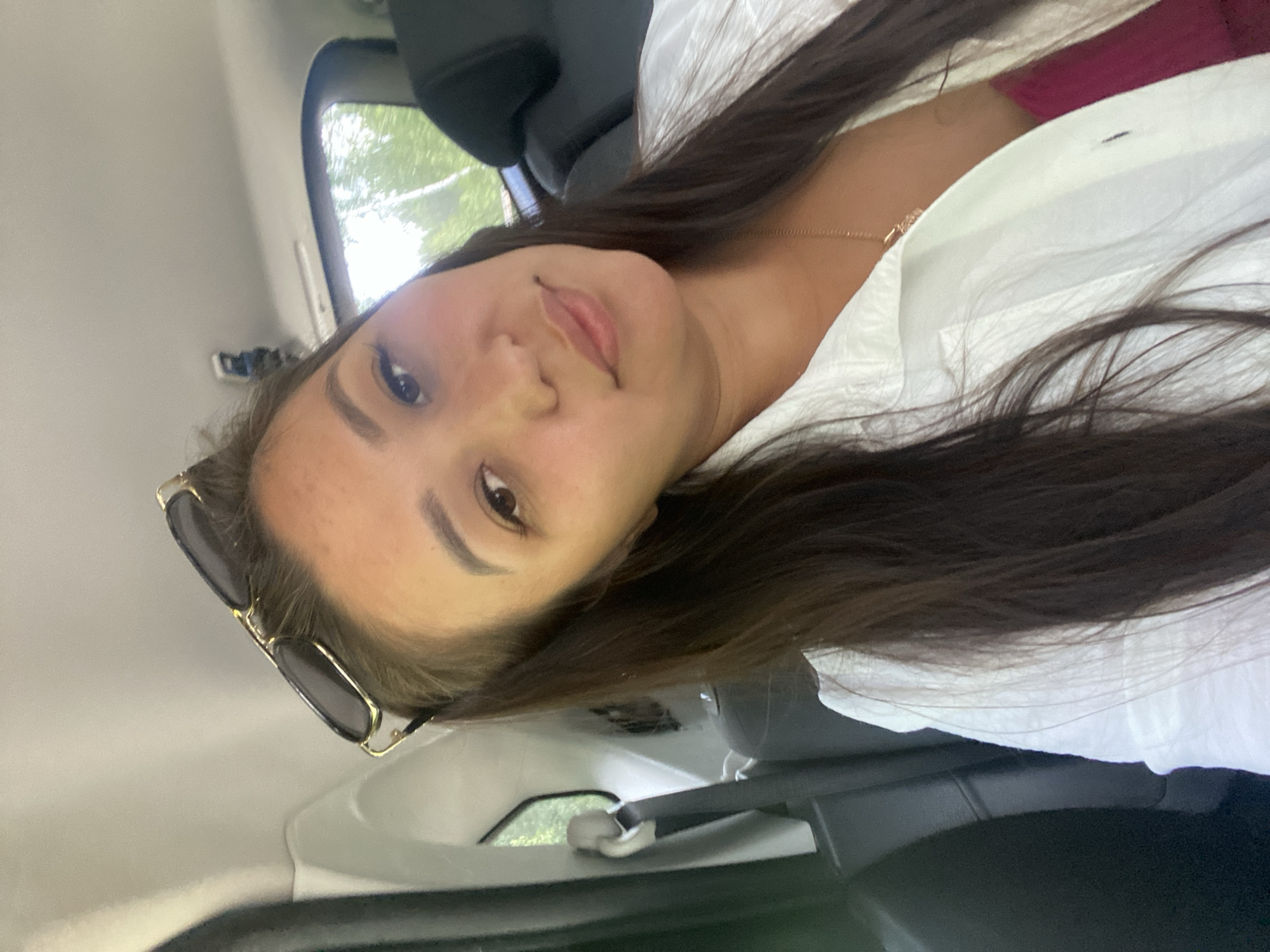A young woman with long dark hair, wearing sunglasses on her head and a white open shirt over a pink top, is taking a selfie in the back seat of a car. The interior of the car is visible, with black seats and a seatbelt. Outside the window, trees and greenery can be seen. The scene appears to be in a rural or natural setting, possibly on a road trip. The image was taken on a sunny day, casting shadows and highlights. The location is identified as Kashkarovo, Russia.