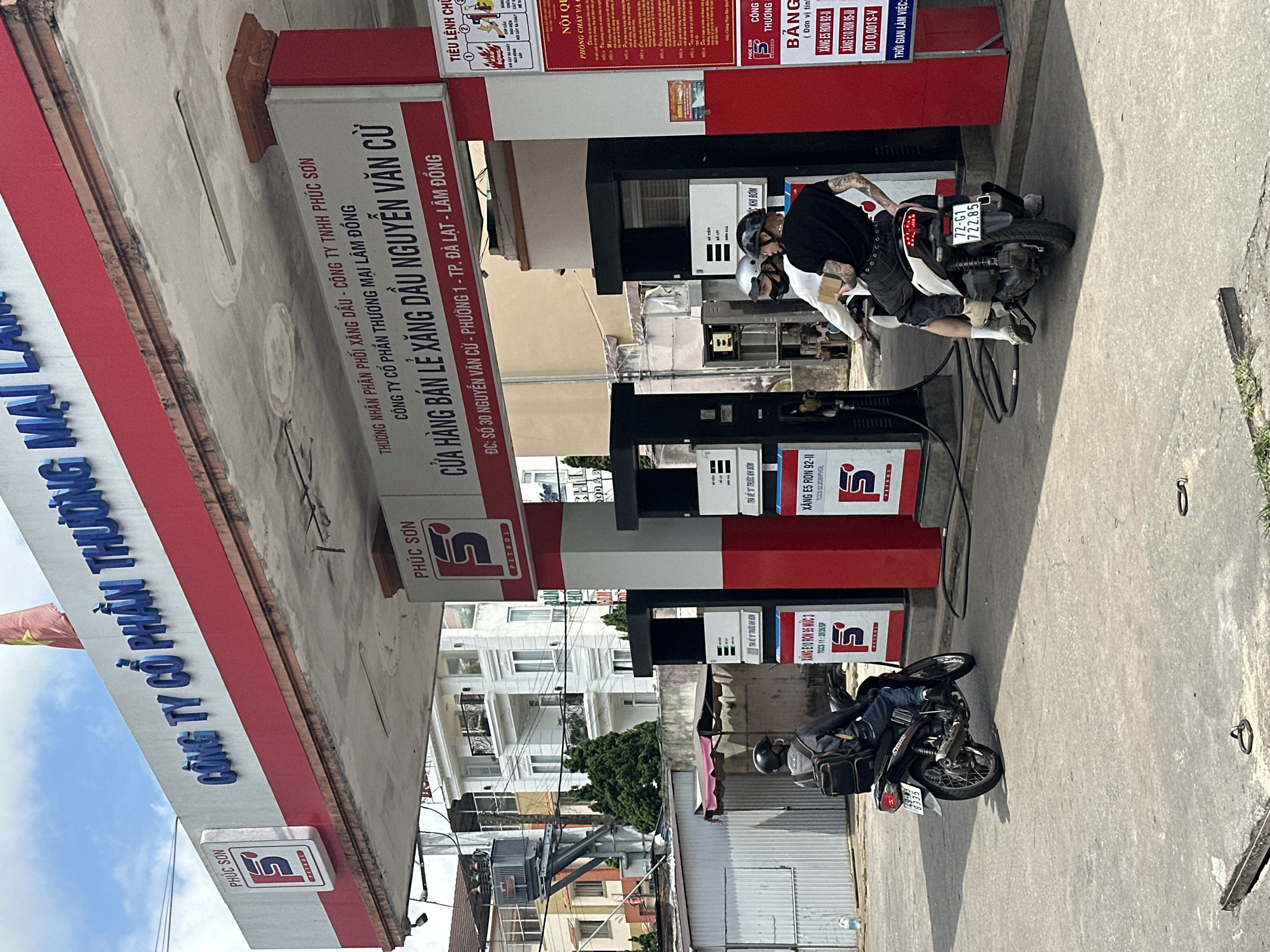 At a "Phuc Son Petrol" gas station in Da Lat, Vietnam, two men on a scooter and one man on a motorcycle are present. The gas station has red and white signage with Vietnamese text indicating the company name and address. The man on the motorcycle is wearing a black jacket and backpack, while the men on the scooter are wearing casual clothing and helmets. The environment appears to be an urban street, with buildings and power lines visible in the background. The weather is overcast.