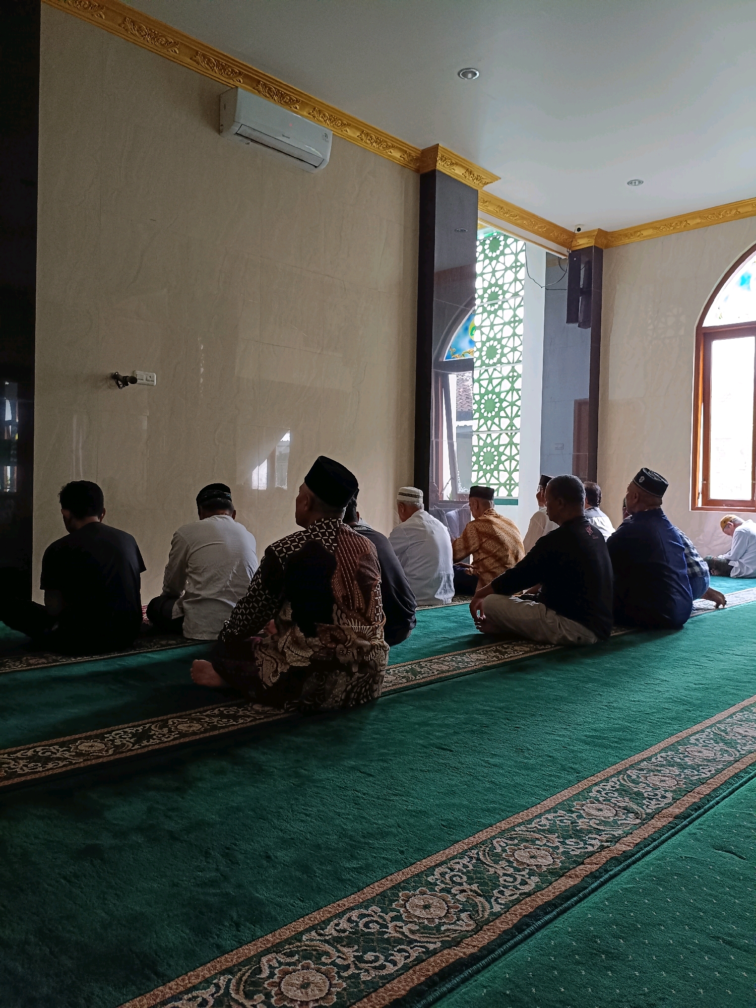 A group of men are seated on a green carpet in a room with beige walls, gold trim along the ceiling, and a large window with decorative glass. Some of the men are wearing traditional Islamic attire, including peci hats and patterned sarongs. One man in the foreground is wearing a black peci and a patterned shirt and sarong. The men are facing away from the camera, seated in rows on the carpet. An air conditioner unit is visible on the wall. The room appears to be a place of worship, possibly a mosque, in Maguwoharjo, Indonesia.