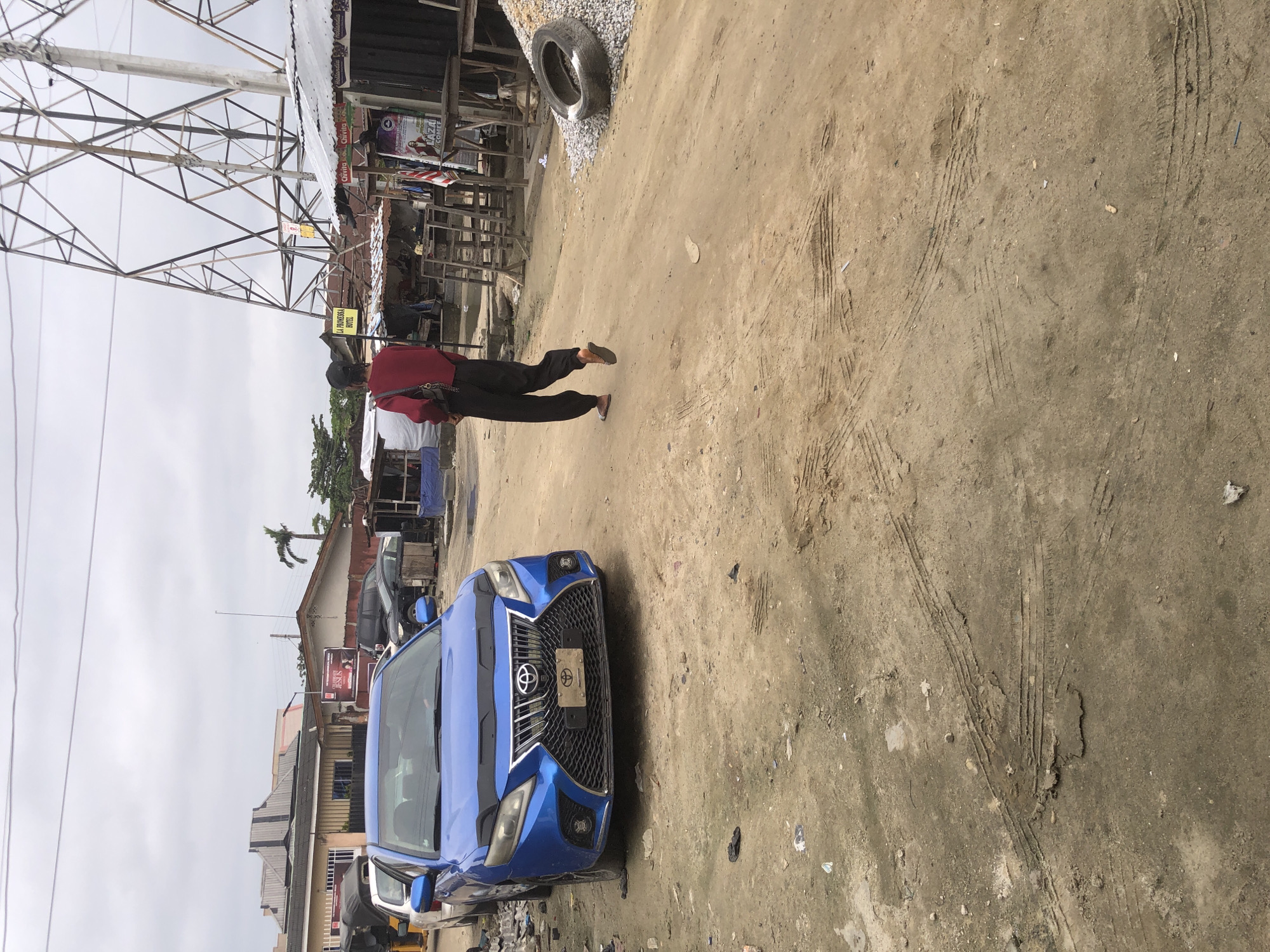 In the city of Ado, Nigeria, an outdoor scene unfolds with a dirt road and a blue sedan in the foreground. The car, a Toyota Camry with a custom Lexus-style grille, is parked on the left side of the road. Behind it, other vehicles are visible, including a white car and a black SUV. A person in a red long-sleeved shirt and black pants is walking away from the camera on the right side of the road, towards a collection of makeshift stalls and structures. These structures, some with corrugated metal roofs, appear to be part of a marketplace or informal business area. Prominent in the background is a large electricity pylon. The ground is dusty and uneven, with tire tracks evident. Debris, including a discarded tire and scattered gravel, is present near the stalls. The sky is overcast.