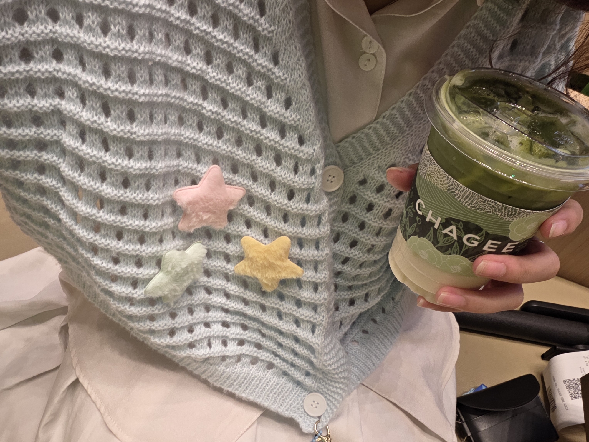 The scene presents a close-up, high-angle shot of a person holding a drink from the chain Chagee. The drink is green with a plastic lid. The person wears a light blue, knitted cardigan with pastel-colored star embellishments. A white collared shirt peeks from beneath the cardigan. A receipt, a dark case, and a black cylindrical object rest on a light brown surface. Given that Chagee is a chain, the exact location within Kuala Lumpur, Malaysia cannot be determined solely from the image.