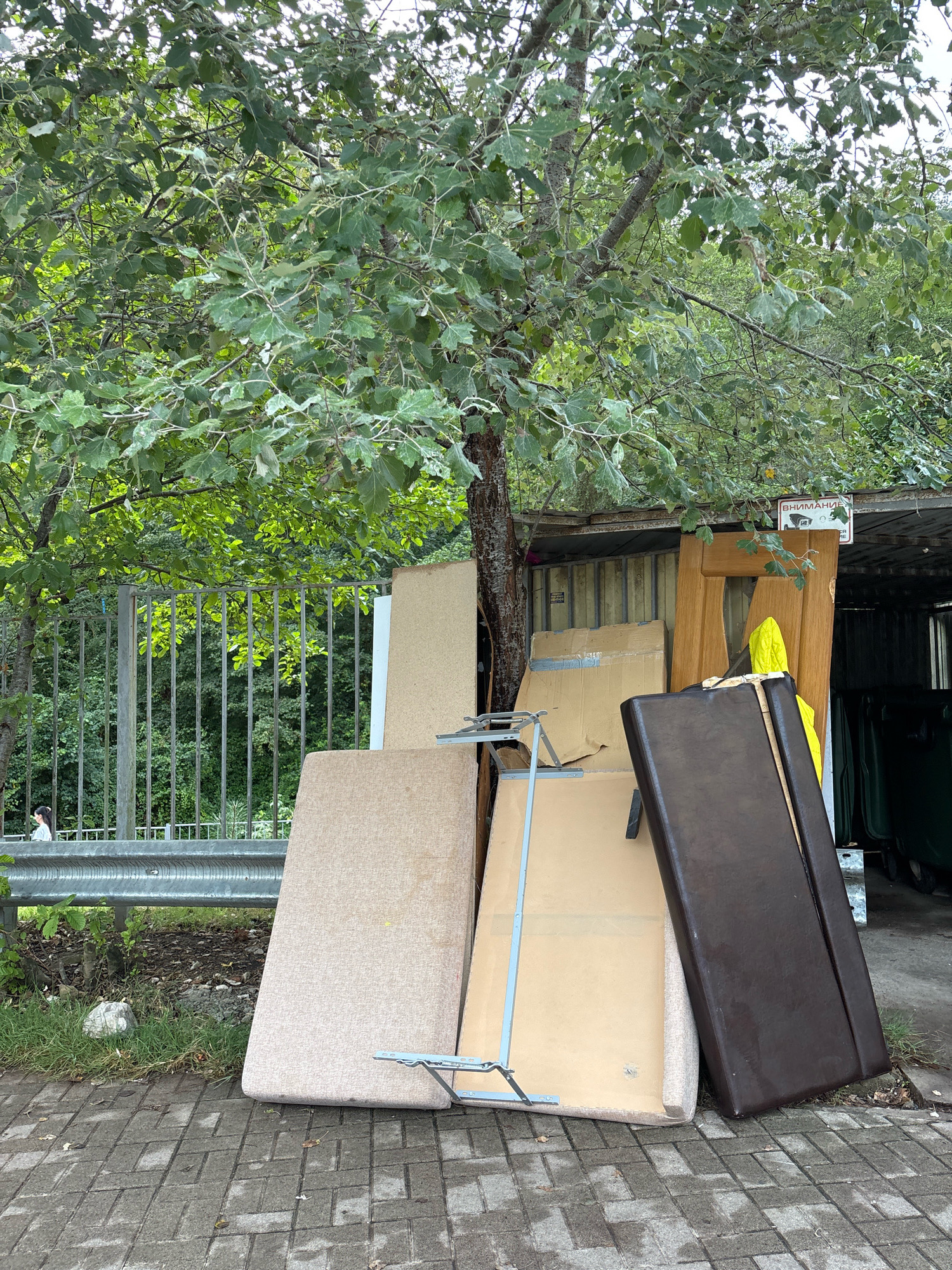 An outdoor image shows a pile of discarded furniture and building materials against a metal structure with a tree in the foreground. The pile includes a brown upholstered sofa cushion, a wooden door, several large flat boards of particleboard or MDF, and a metal folding frame. Behind the pile, a corrugated metal wall is visible, and further back, green refuse bins are partially obscured. To the left of the pile, a metal fence runs parallel to a paved walkway. Beyond the fence, a dense area of trees and foliage forms the background. A single person is visible in the distance on the left side of the image, walking along a path beyond the fence. The ground in the foreground is paved with grey rectangular bricks. A sign with Cyrillic text and a camera icon is mounted on the metal structure above the door. The scene appears to be a collection point for bulky waste in an outdoor urban setting, possibly in Sochi, Russia, given the presence of Cyrillic script. The overall atmosphere is one of disarray and disposal.