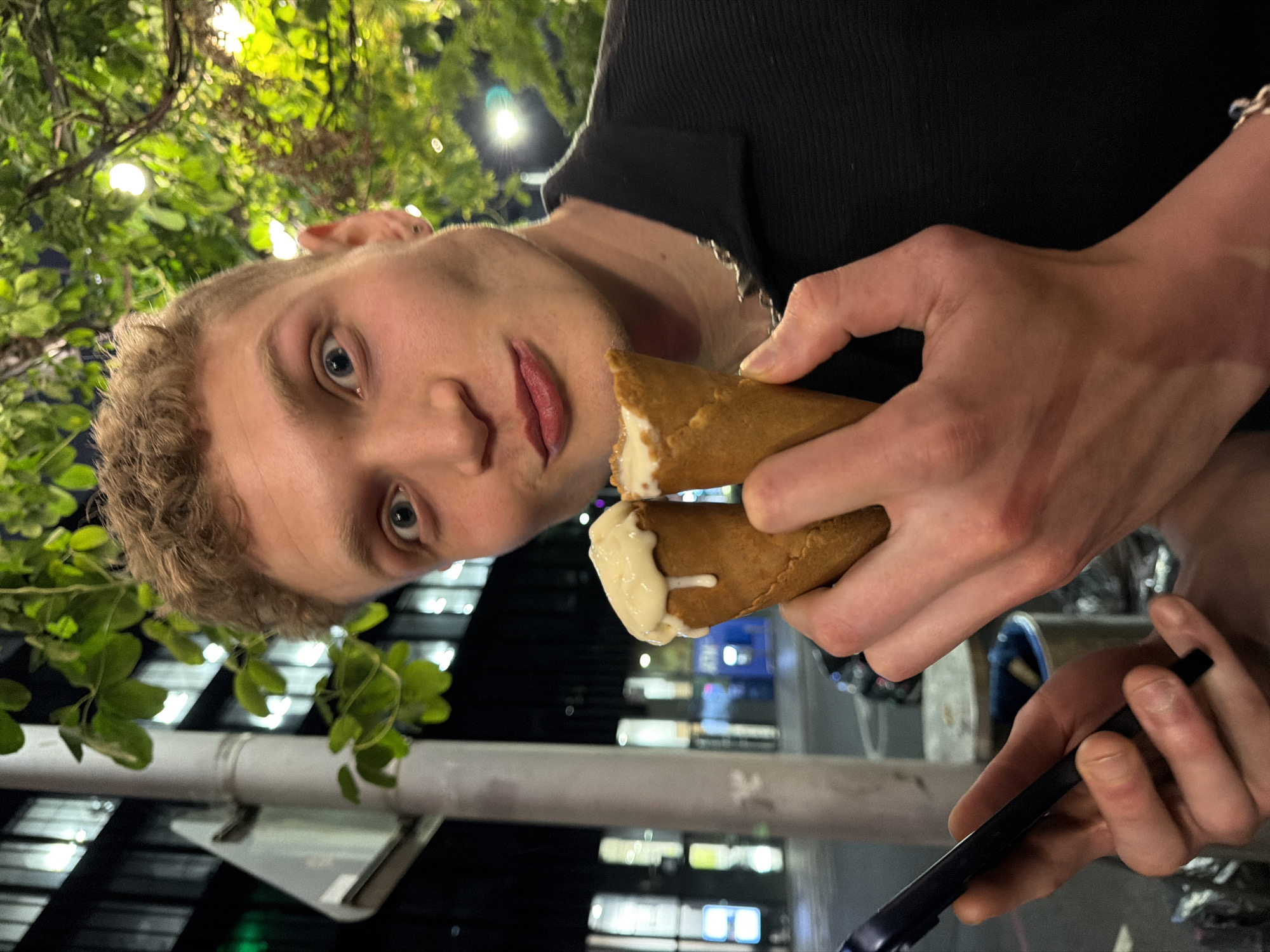 A young man is captured in a close-up, low-angle portrait, taken at night in an urban outdoor setting in London, United Kingdom. He has short, light brown hair, fair skin, and striking blue eyes, gazing directly into the camera with a neutral, slightly inquisitive expression. He wears a black, ribbed collared shirt and a silver chain around his neck.
The man is actively eating or about to eat a dessert. He holds two pieces of a light brown, conical pastry, resembling a waffle cone or crepe, which are filled with a white, creamy substance, likely ice cream or another sweet cream. One piece is positioned near his mouth, while the other piece below it shows some of the white cream visibly dripping. In his other hand, a dark, slender object, possibly a phone or a vape pen, is visible, and he wears a bracelet on his wrist.
The background is dark, indicating nighttime. Lush green foliage from a plant or tree is prominent in the foreground and overhead, suggesting an area with natural elements amidst the city. Blurred, illuminated windows and lights from distant buildings create an urban backdrop, with a grey horizontal beam or pipe also visible. No visible text is present in the image.