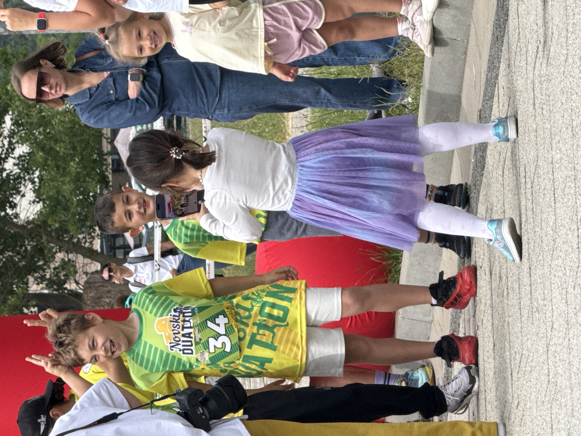 A group of people stand on a light gray tiled or concrete surface. A young girl in a white long-sleeved shirt and purple tutu takes a photo with a smartphone. Next to her, a young boy in a green and yellow patterned shirt poses with hand gestures. An adult woman wearing sunglasses stands behind them. In the background are green trees and a building. The setting is Igalo, Montenegro.