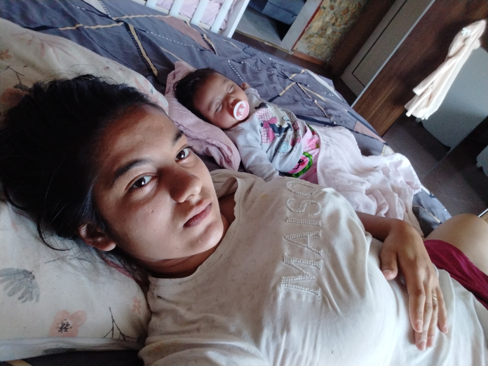 A woman lies on a bed, looking at the camera. Next to her, a baby sleeps with a pacifier in its mouth. The location is Unknown, Unknown. The woman is wearing a white shirt. The baby is wearing a patterned shirt and pants. The bed has a grey and blue patterned blanket. A white crib is visible in the background.