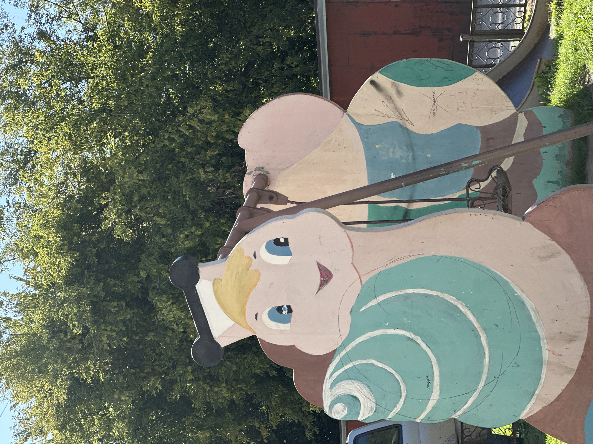 The image depicts a brightly painted playground structure, shaped like a cartoonish snail. The snail has a large, round, light-pink head with blue eyes, a wide smile, and blonde hair. It wears a black and white hat. Its body is designed to resemble a spiral shell, painted in shades of light teal and white. The structure appears to be made of wood or a similar material and is weathered, showing signs of age and possibly graffiti. Behind the snail, a metal swing set pole is visible, and to the right, a section of a metal slide with a blue chute can be seen. The background consists of dense green foliage from trees, a red building, and a clear blue sky. The ground is covered with green grass. There are no people or animals visible in the image. The overall setting suggests an outdoor public space, likely a park or a kindergarten playground in the city of Biysk, Russia, given the context.