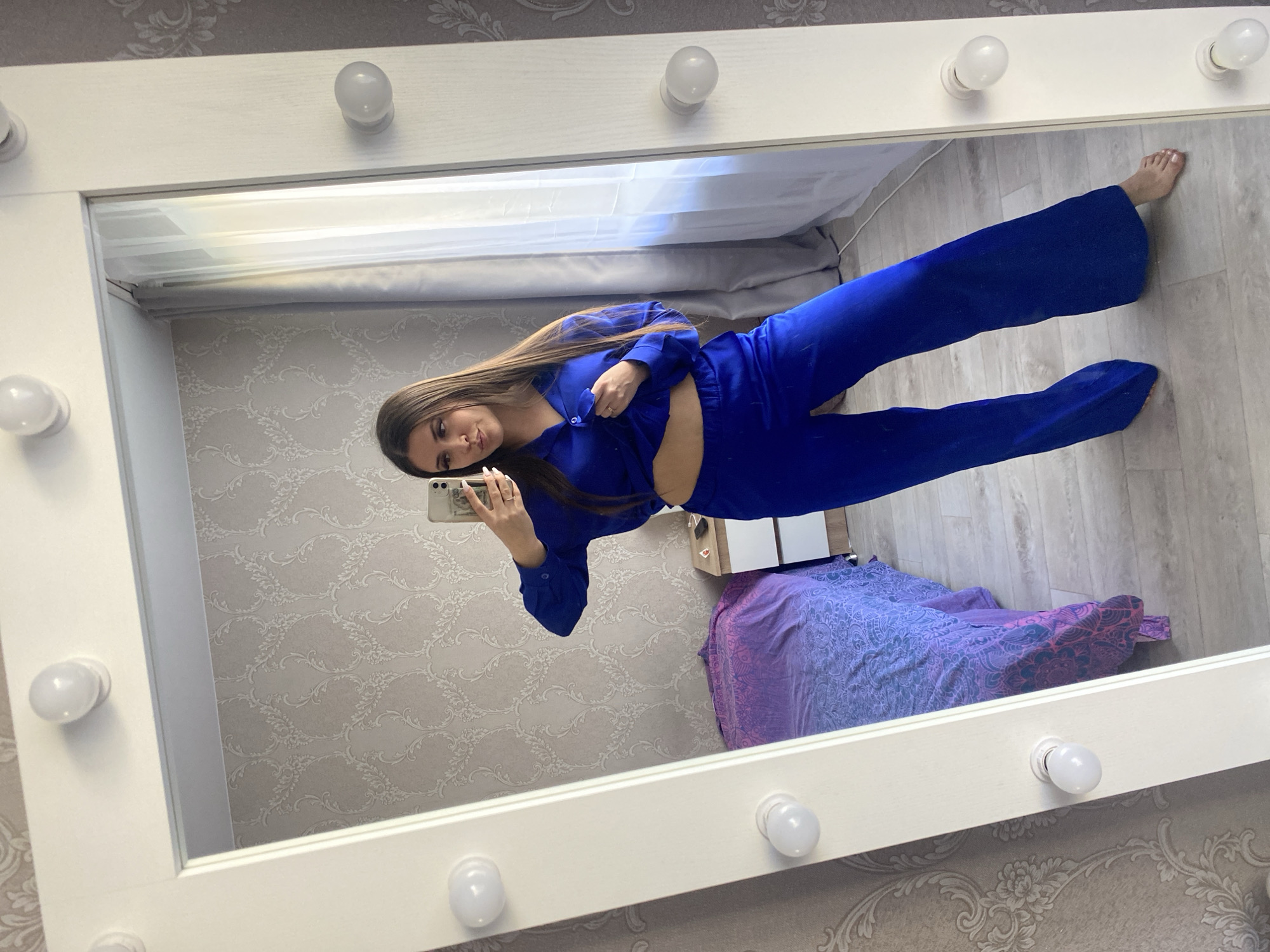 A woman is standing in front of a mirror taking a selfie with her smartphone. She is wearing a two-piece blue outfit. The mirror has light bulbs around the perimeter and is reflecting a room with patterned wallpaper, a bed with a purple and blue patterned comforter, and a small white cabinet. The location is unknown and there is no indication of this image being taken in Chaikovsky, Russia.