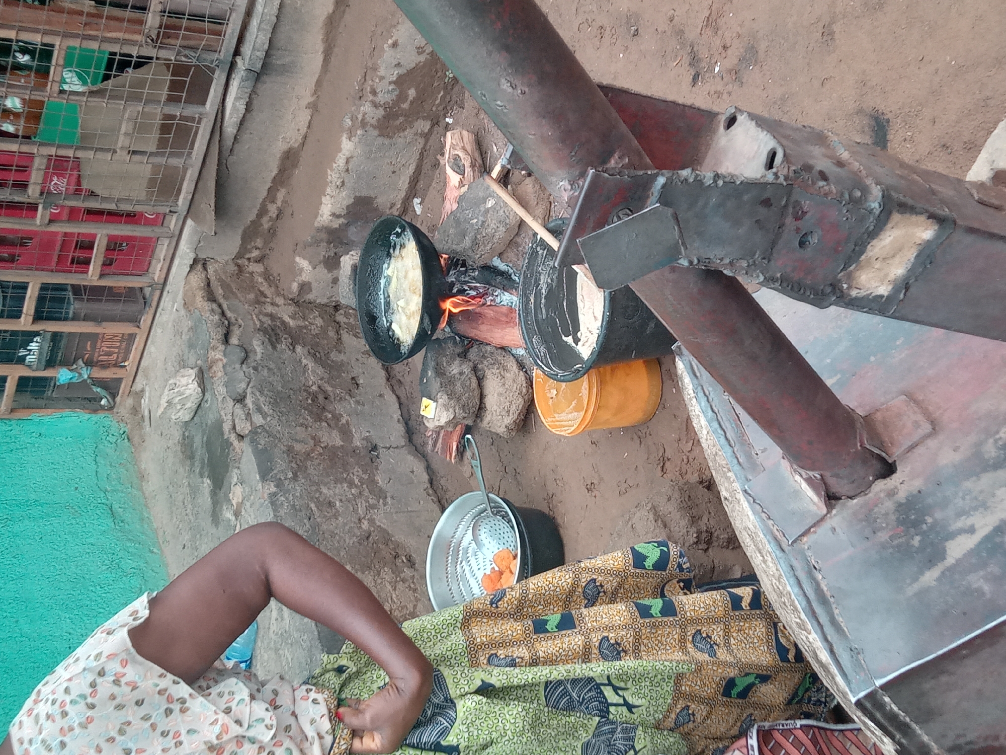 In Sabon Tasha, Nigeria, a woman wearing a floral patterned top and a patterned green and yellow cloth skirt sits beside a makeshift outdoor cooking area. The cooking apparatus consists of a metal structure supported by pipes, with a fire burning underneath two black pots. One pot contains bubbling oil with food frying, while the other appears to hold uncooked batter. A metal strainer basket filled with cooked, orange-colored food items sits nearby. The ground is bare dirt. A building with a turquoise wall and a cage-like structure filled with items is visible in the background.